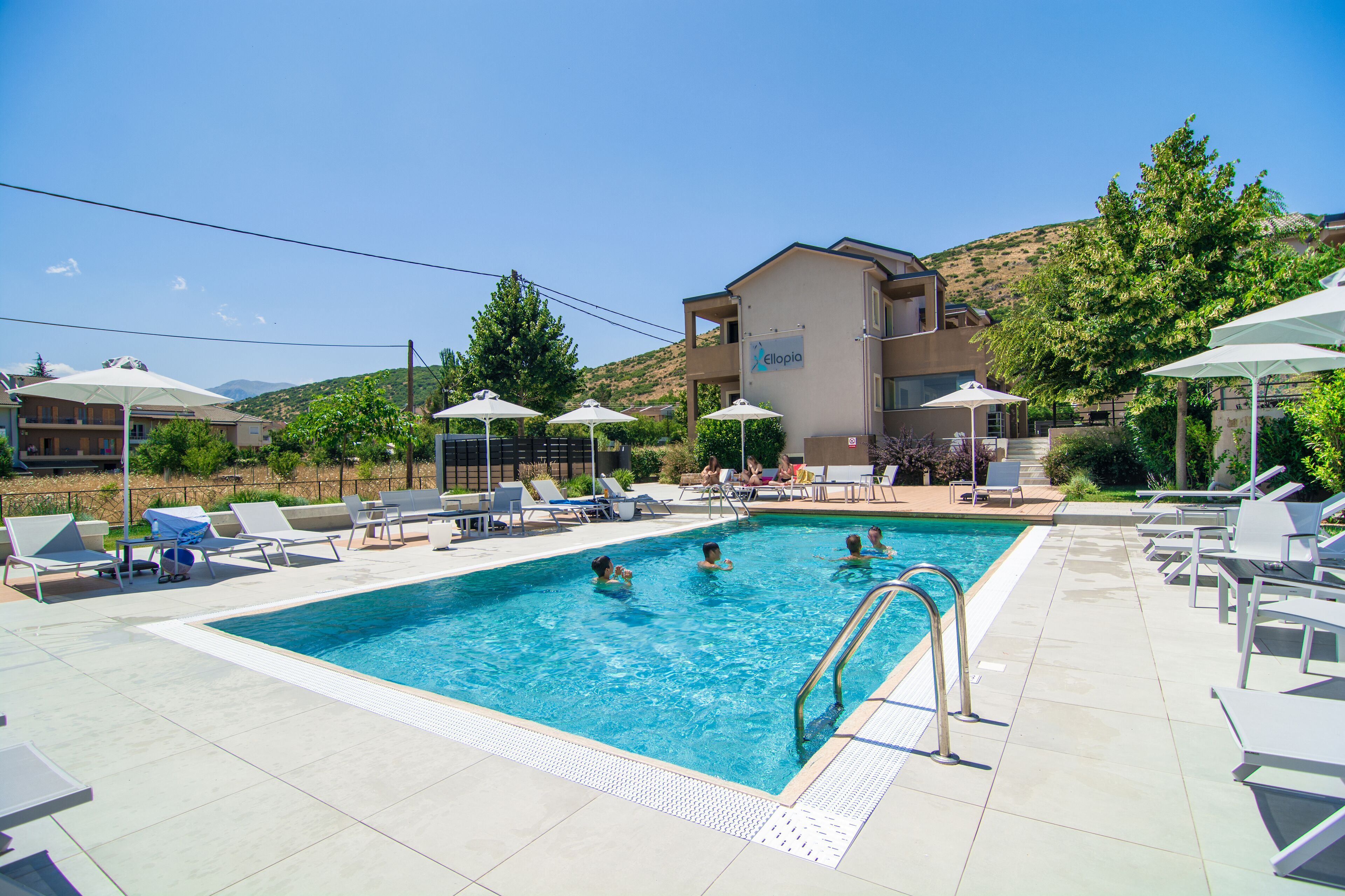 Piscina con sillas de descanso y sombrillas, rodeada de edificios y árboles.