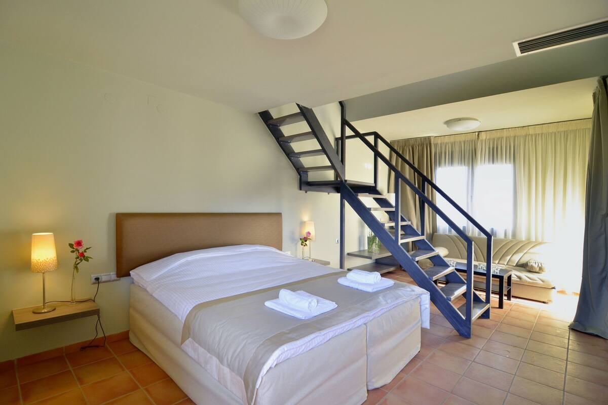 Habitación de hotel con cama, mesita de noche con lámpara, una flor, una escalera y una zona de estar con sofá y cortinas.