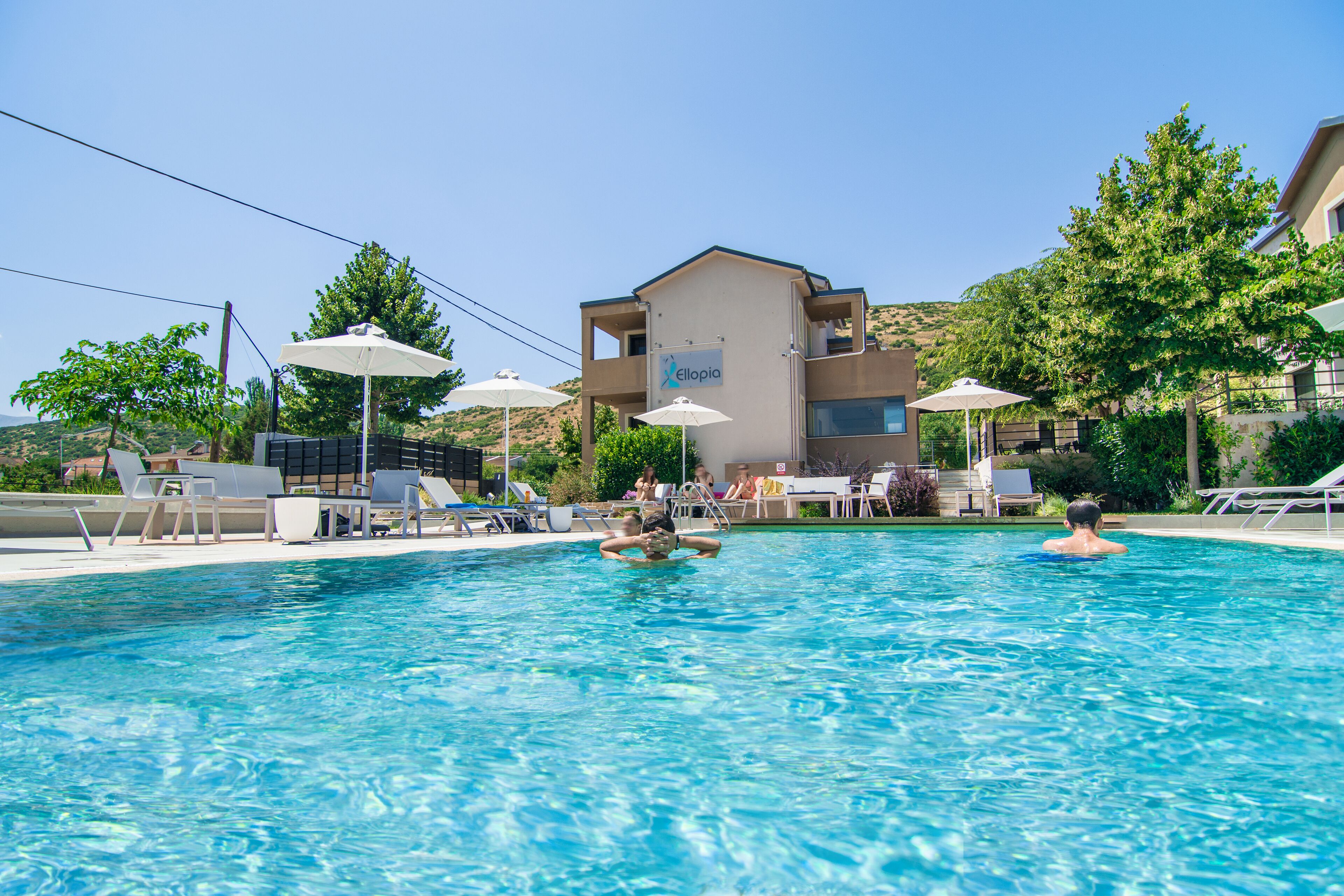Una piscina con sillones reclinables y sombrillas, un edificio con la señalización 'ellopia' y un paisaje montañoso al fondo.
