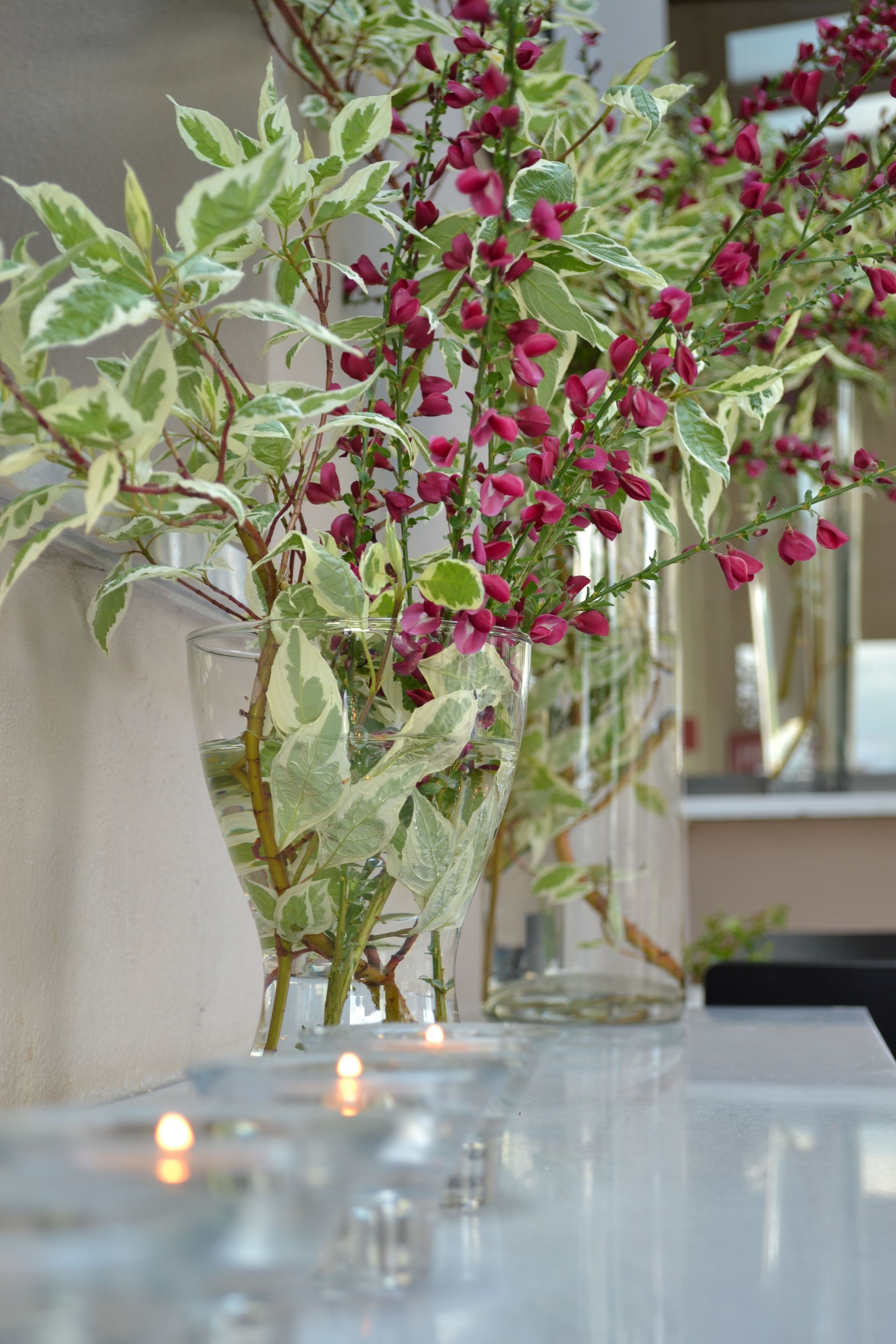Una fila de jarrones de vidrio transparente con hojas verde y blanca y flores rosadas, colocados sobre una mesa con velas.