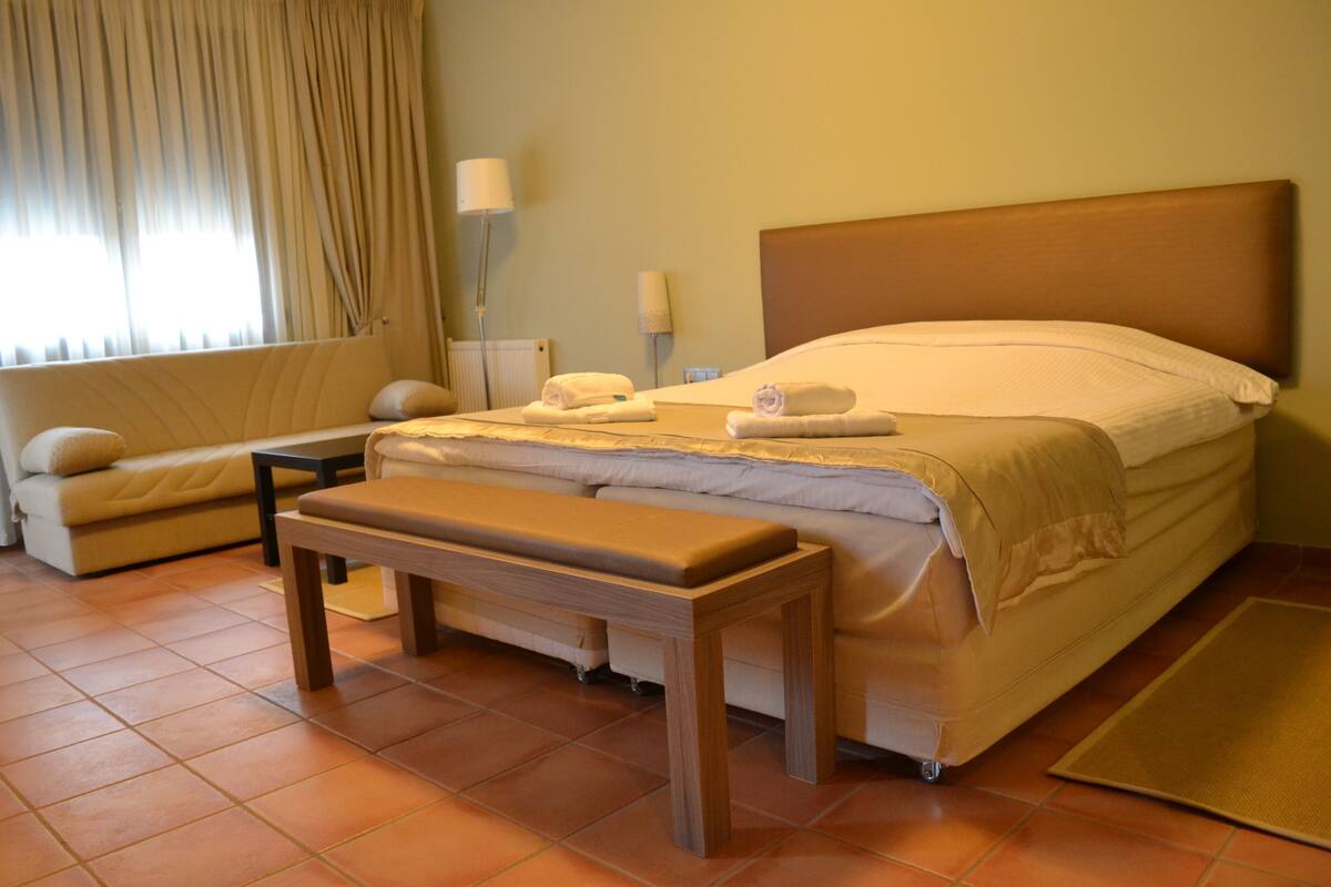 Una habitación de hotel con cama, reposabajares, una mesita, un sofá y una ventana con cortinas.