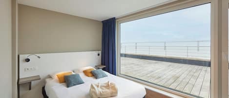 Et moderne hotelværelse med en stor seng, et natbord, og udsigt til en træterrasse gennem et stort vindue.