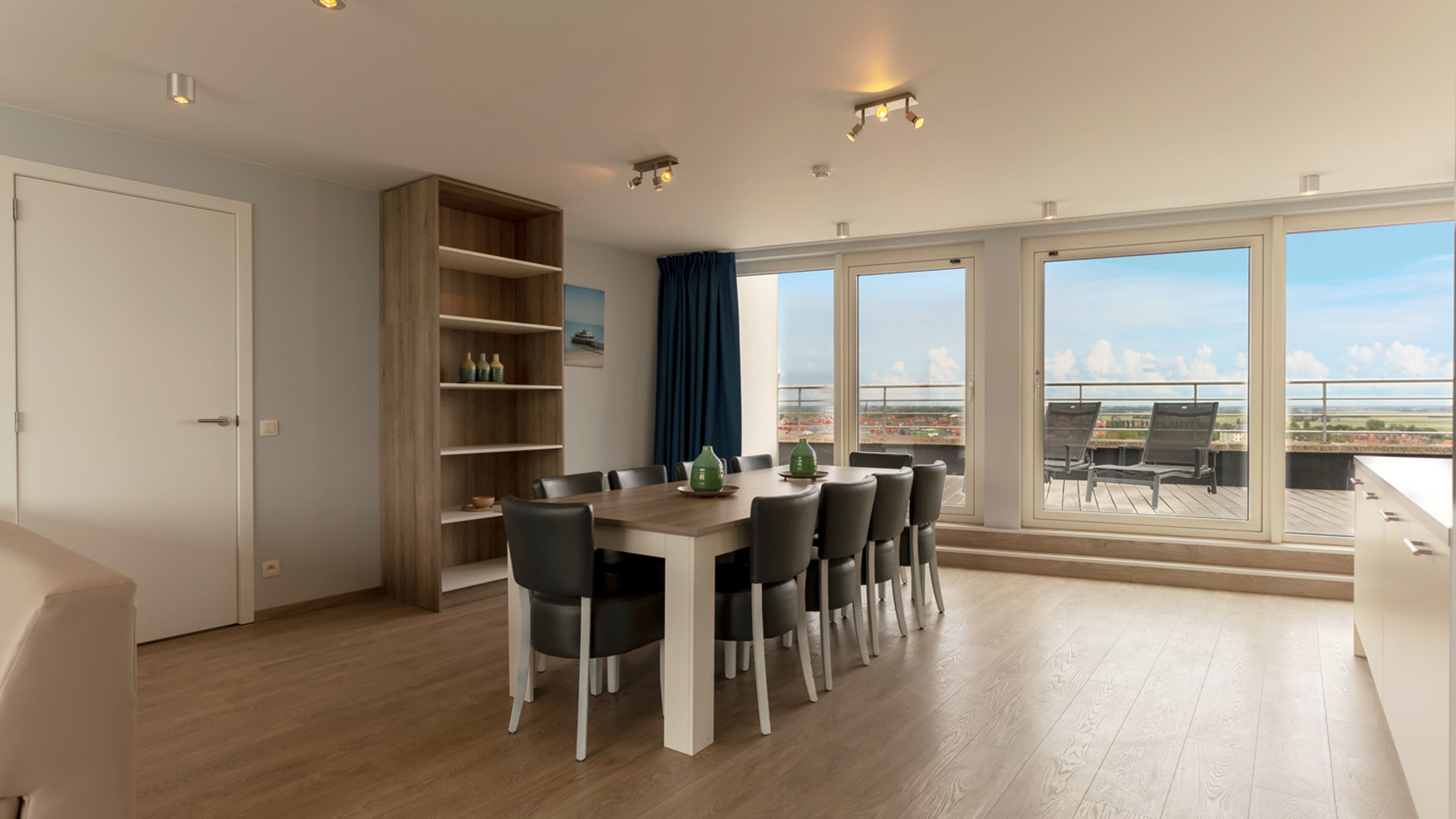 A modern dining area with a wooden table and chairs, a built-in bookshelf, and large windows offering a view of the city.