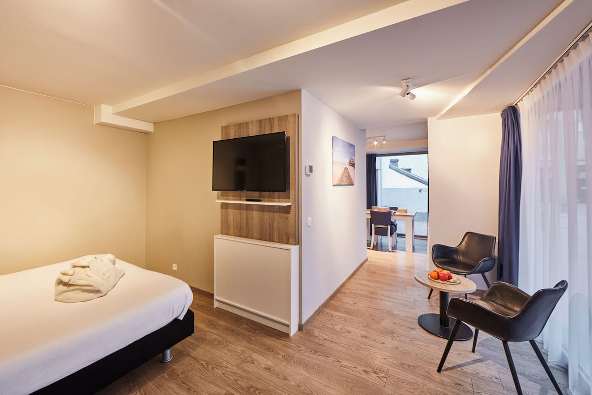 Una habitación de hotel moderna con una cama, un televisor montado en la pared, una pequeña mesa con fruta y una zona de estar con sillas.