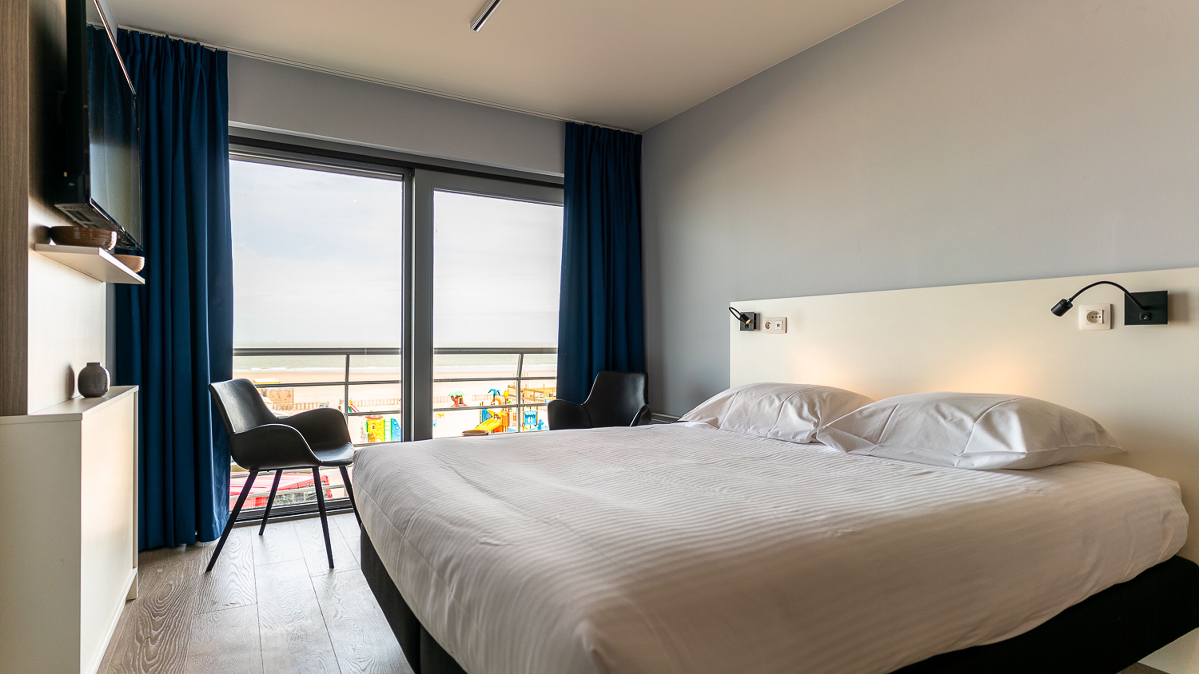 Een hotelkamer met een groot bed, een stoel, een bureau en uitzicht op het strand.