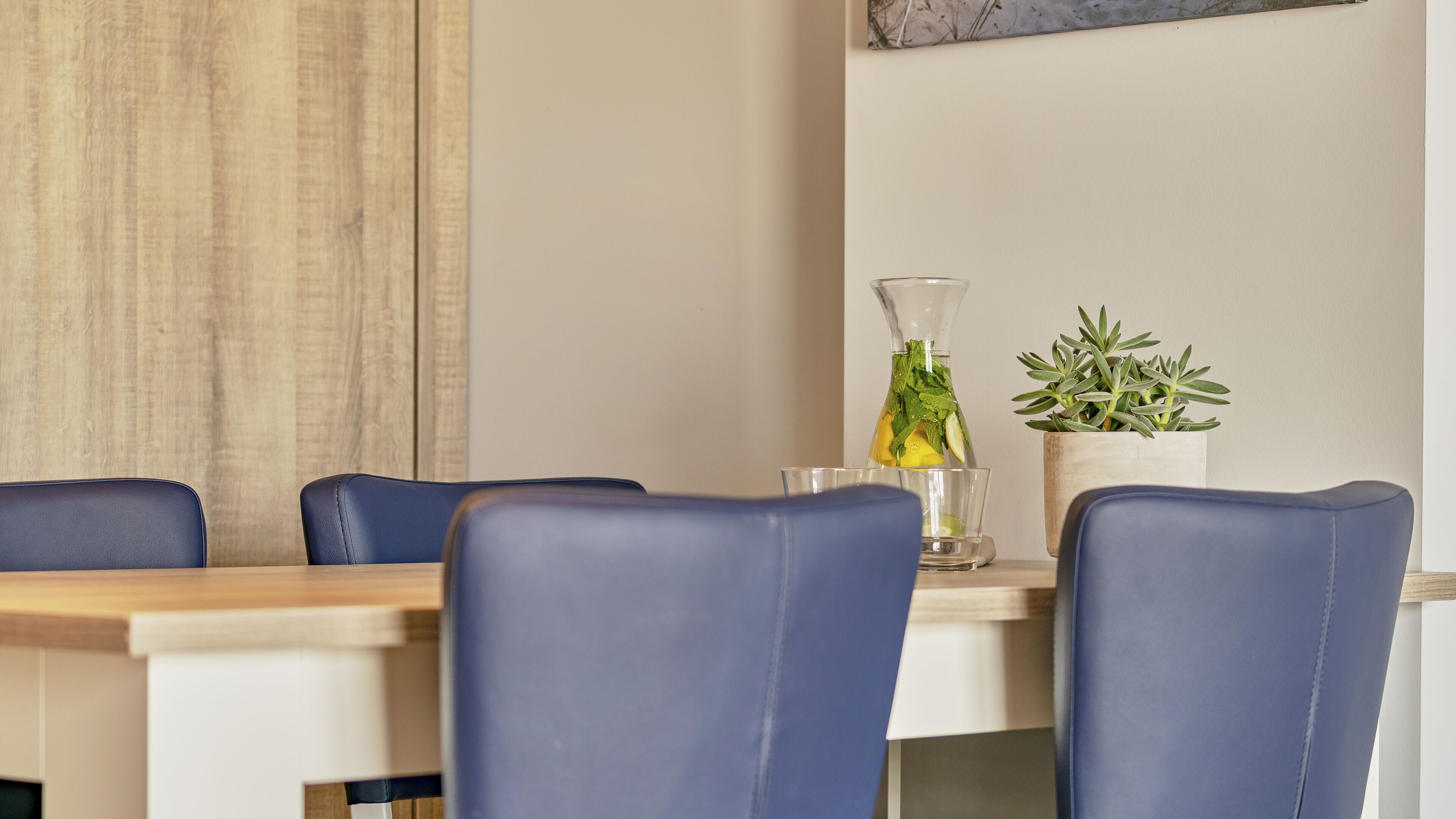 Un coin repas avec une table en bois et des chaises bleues. On peut voir une bouteille en verre transparent contenant une plante et un verre posés sur la table.