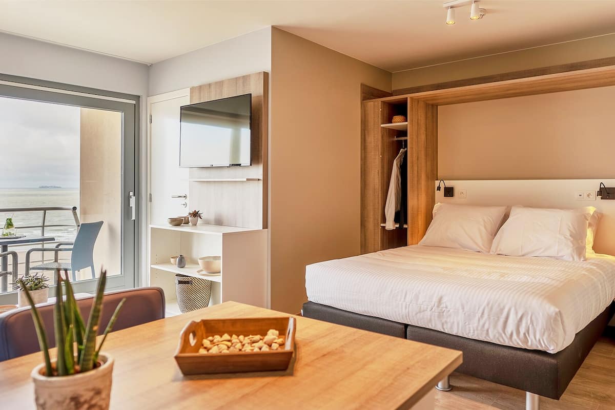Una habitación de hotel moderna con cama, una mesa de comedor con una planta y vista al mar a través de un ventanal grande.