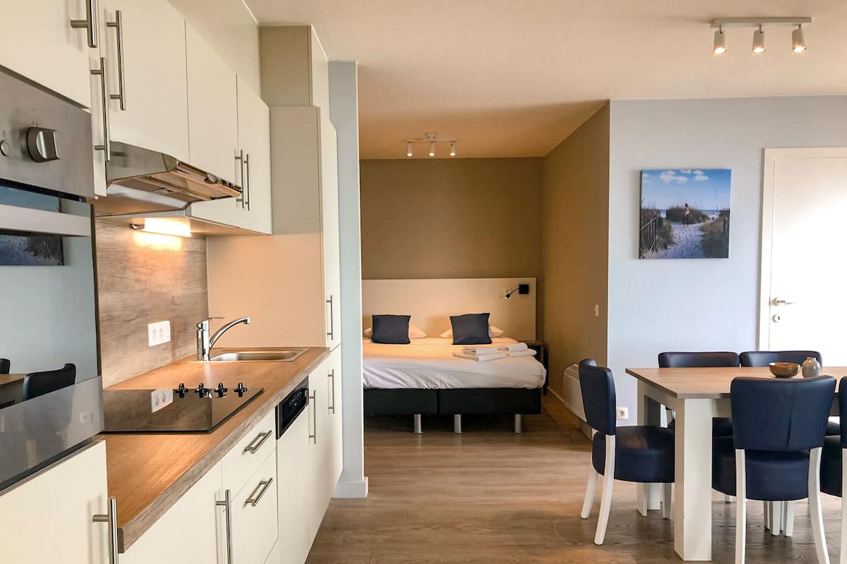 Una cocina moderna con estufa integrada, una zona de comedor con mesa y sillas de madera, y un dormitorio con cama y mesitas de noche.