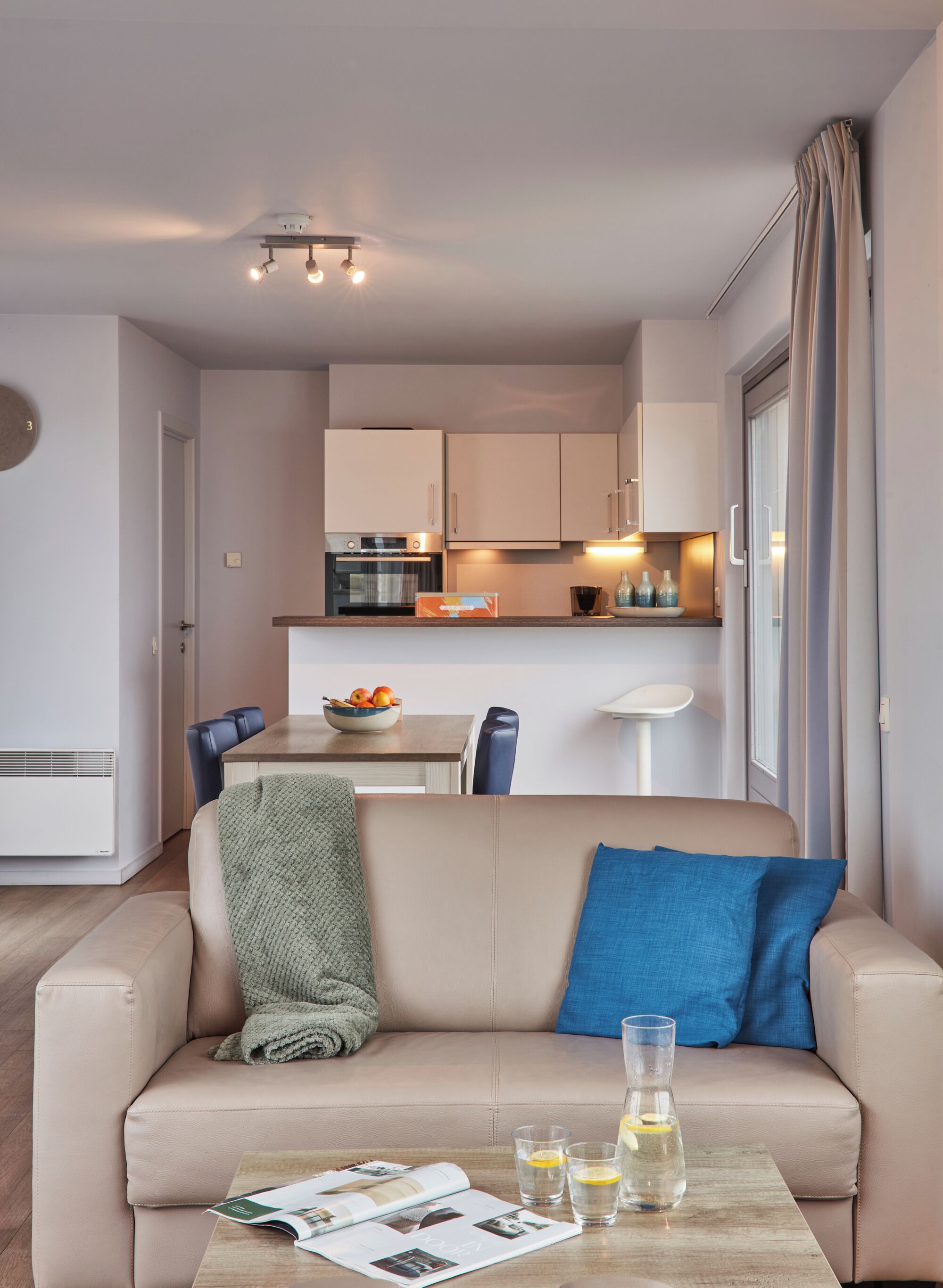 Uma sala de estar moderna com um sofá bege, uma mesa de centro com revistas e copos, uma área de jantar com uma tigela de frutas e uma cozinha com armários brancos e um forno embutido.