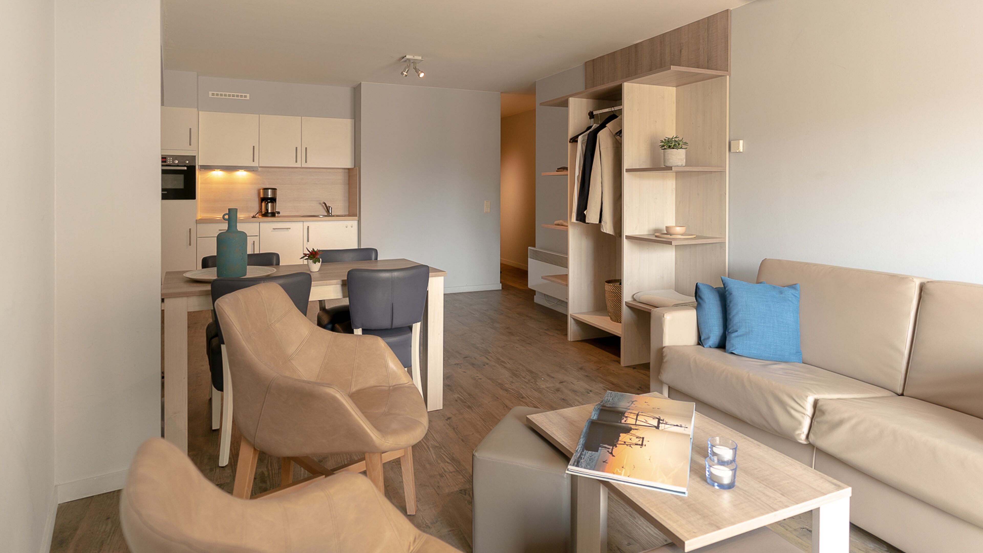 Een moderne woonkamer met een bank, stoelen, een salontafel en een keukenhoek met kasten en apparatuur.