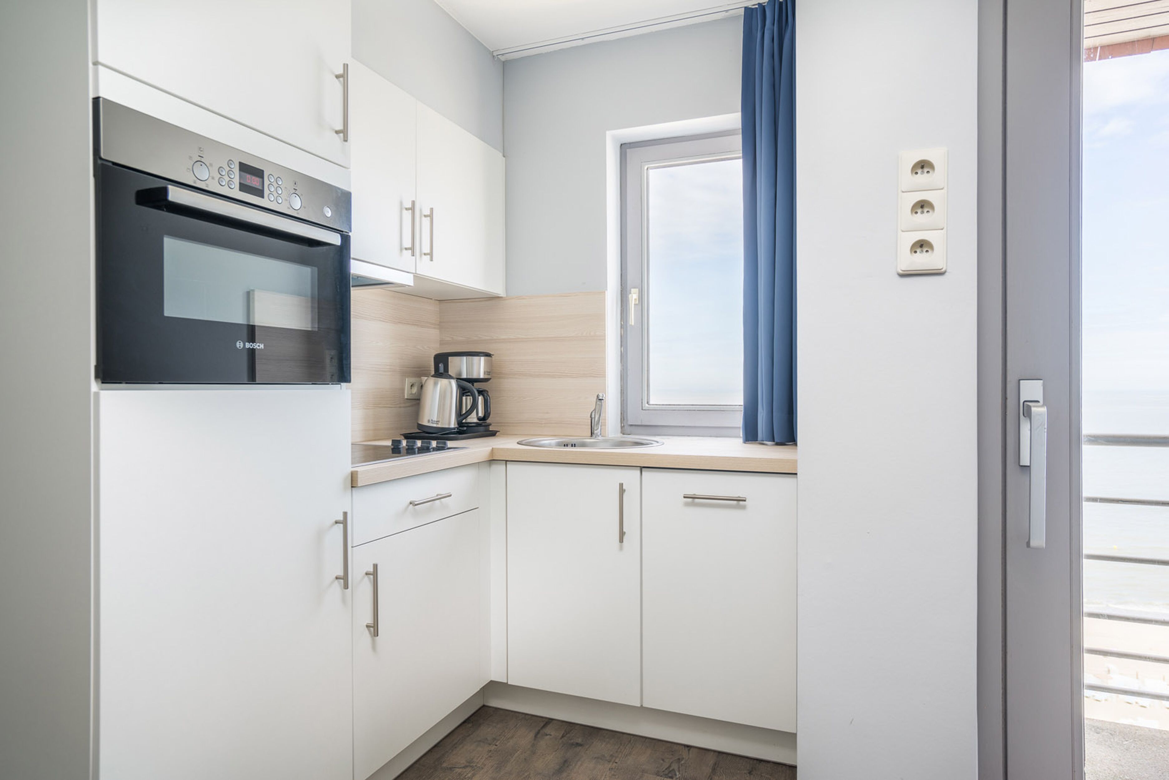A compact kitchen with white cabinets, a Bosch oven, a coffee maker, and a window with blue curtains.