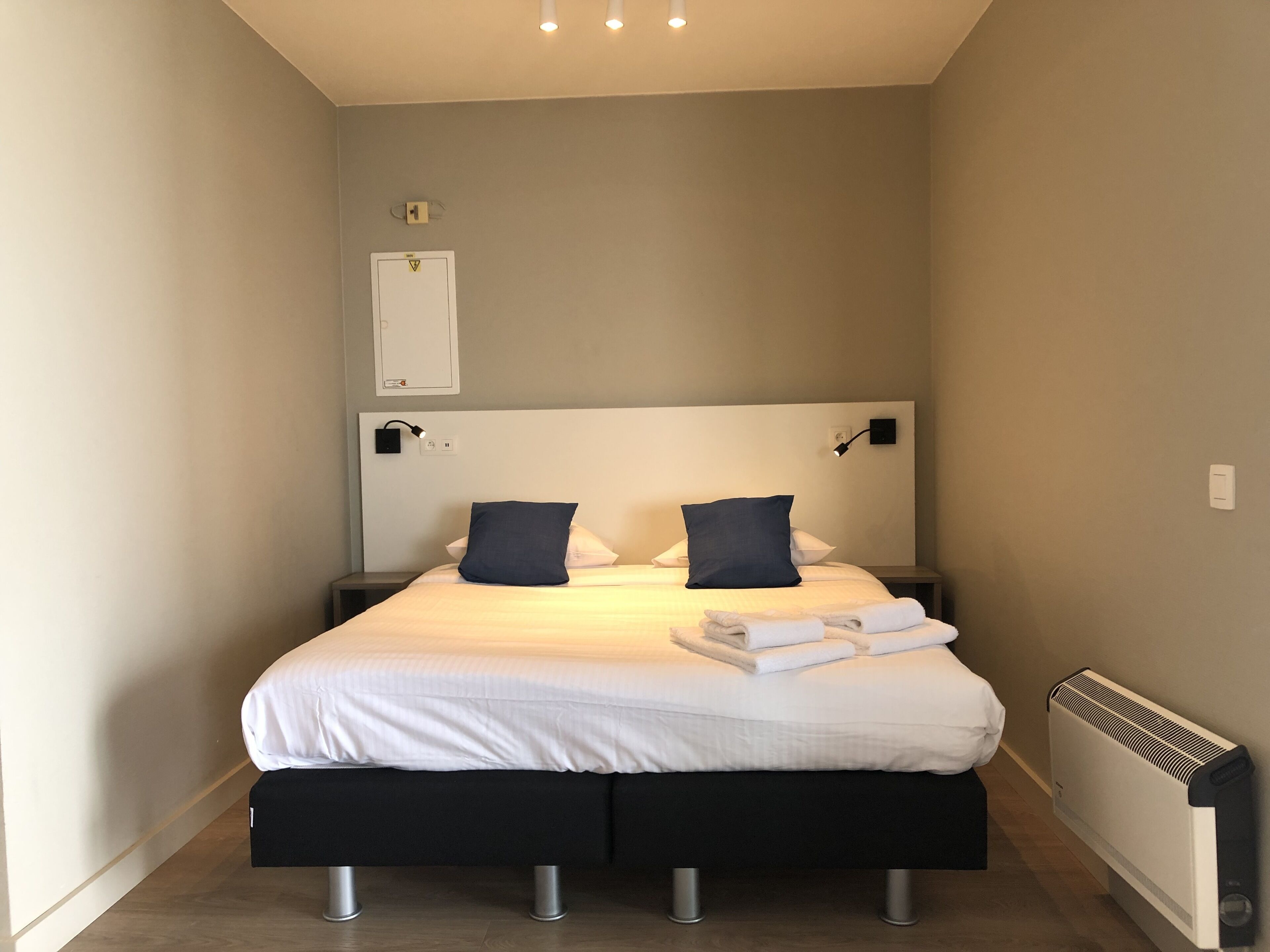 A narrow hotel room with a bed, two bedside tables, a wall-mounted light, a radiator, and a small shelf.