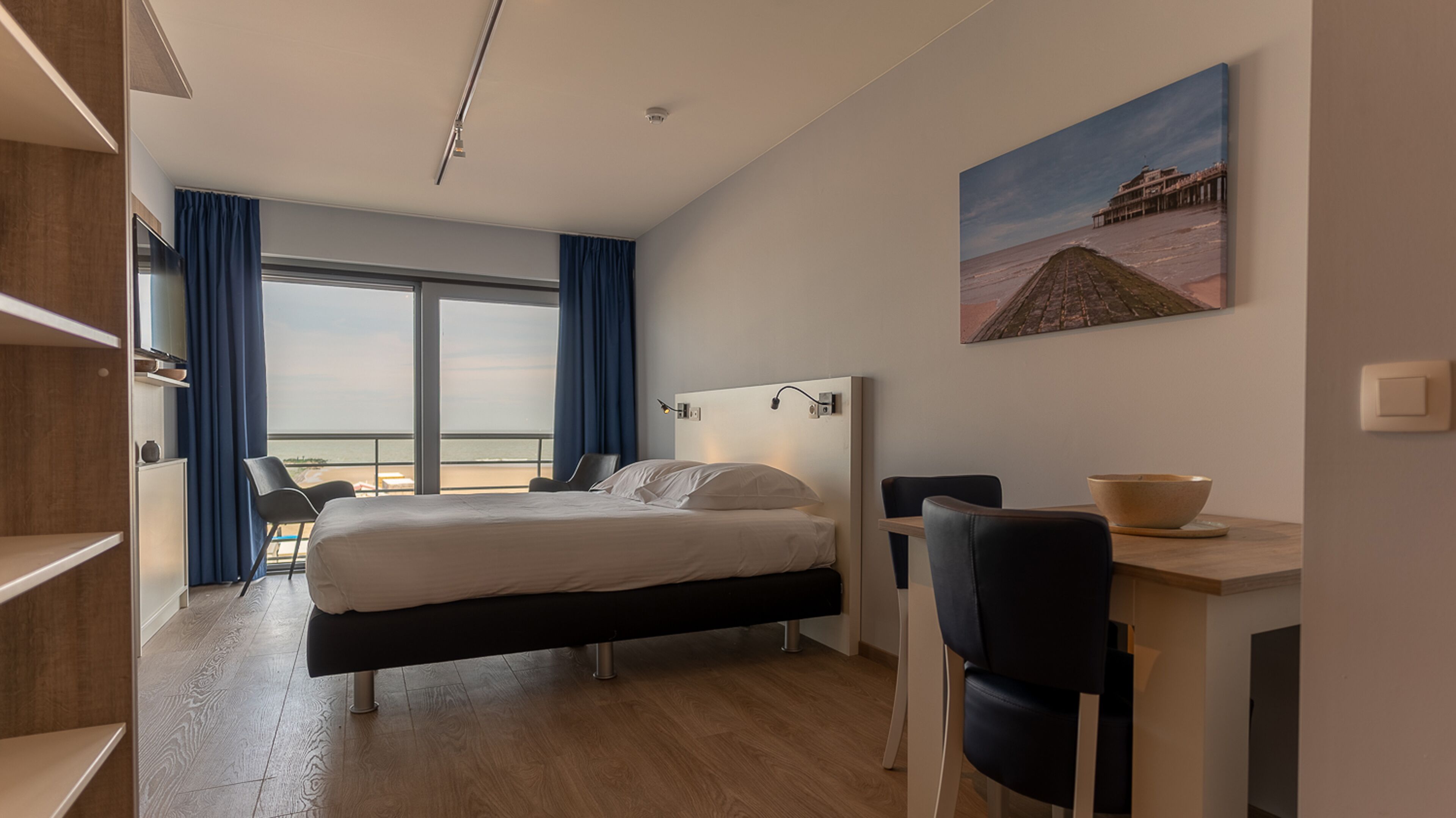A modern hotel room with a bed, a desk, a chair, and a large window offering a view of the beach.