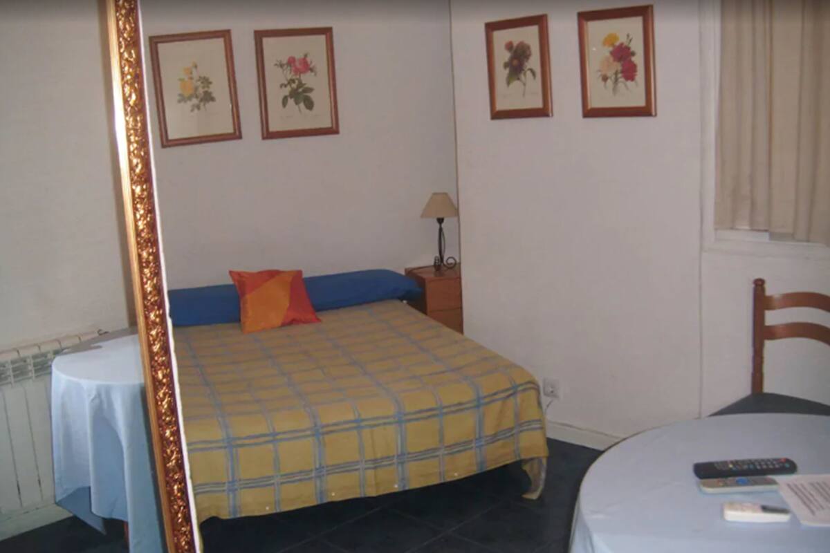 A bedroom with a bed, a nightstand, a lamp, a chair, and a table with a remote control and a book.