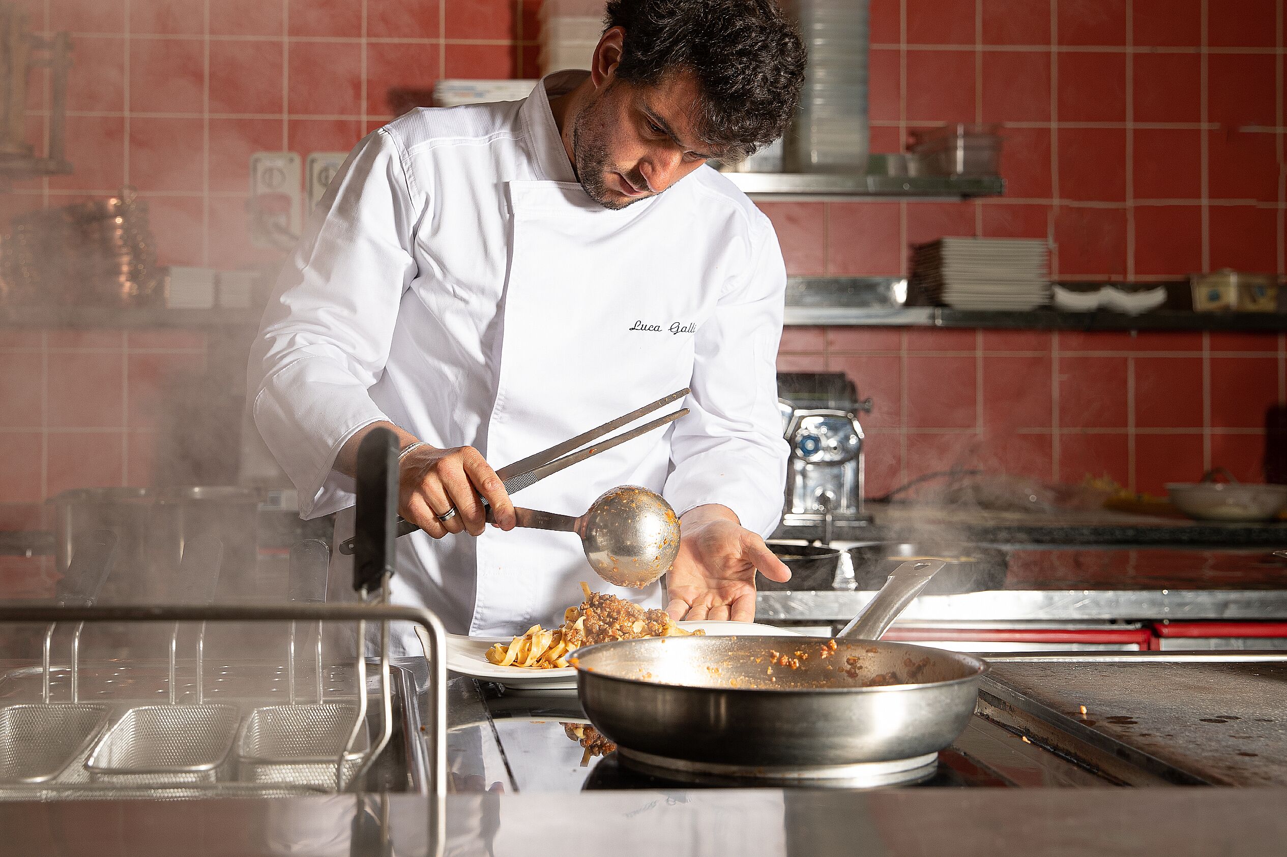 A chef in a white uniform with "Lucca Galli" on the apron, cooking in a kitchen with red tiled walls and stainless steel equipment.