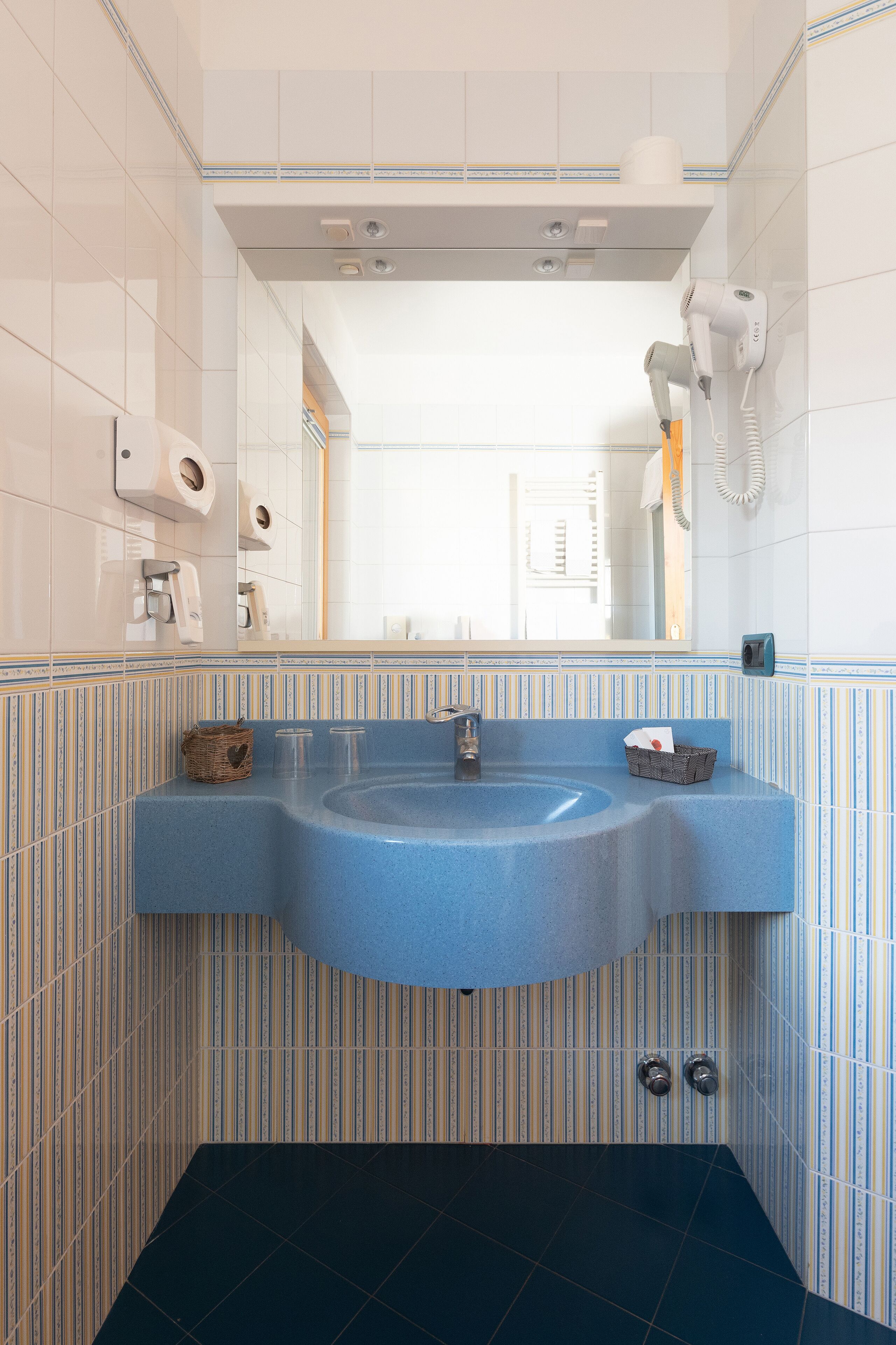 A bathroom with a blue sink, striped wall tiles, a mirror, and a hairdryer on the wall.