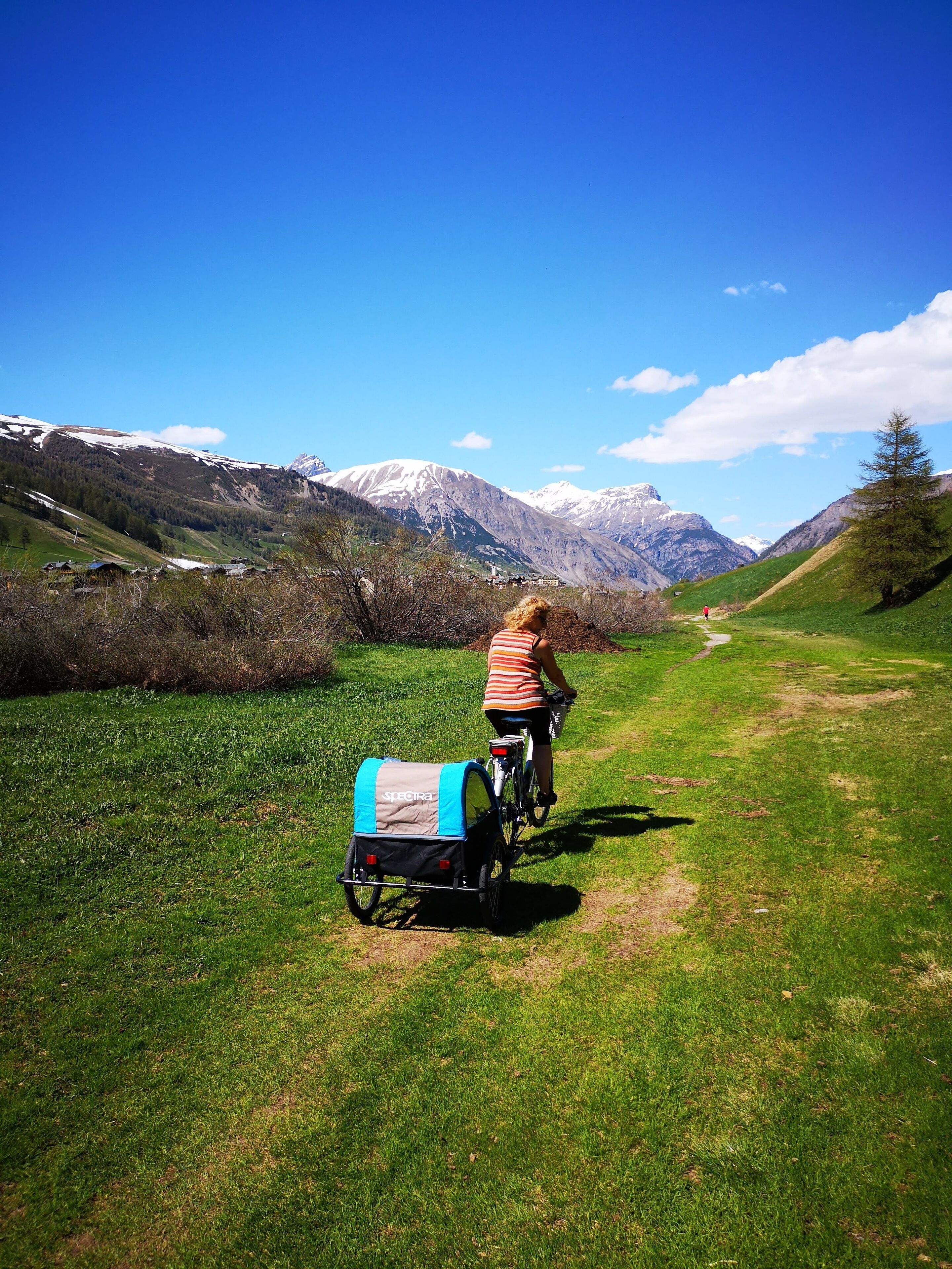 A person riding a bicycle on a grassy path with a blue and black bag attached to the back.