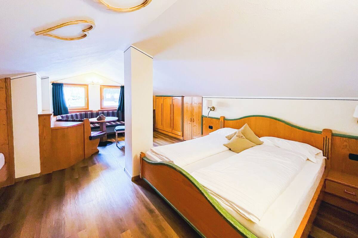 A hotel room with a bed, wooden wardrobe, a small seating area with a table, and a window with curtains.