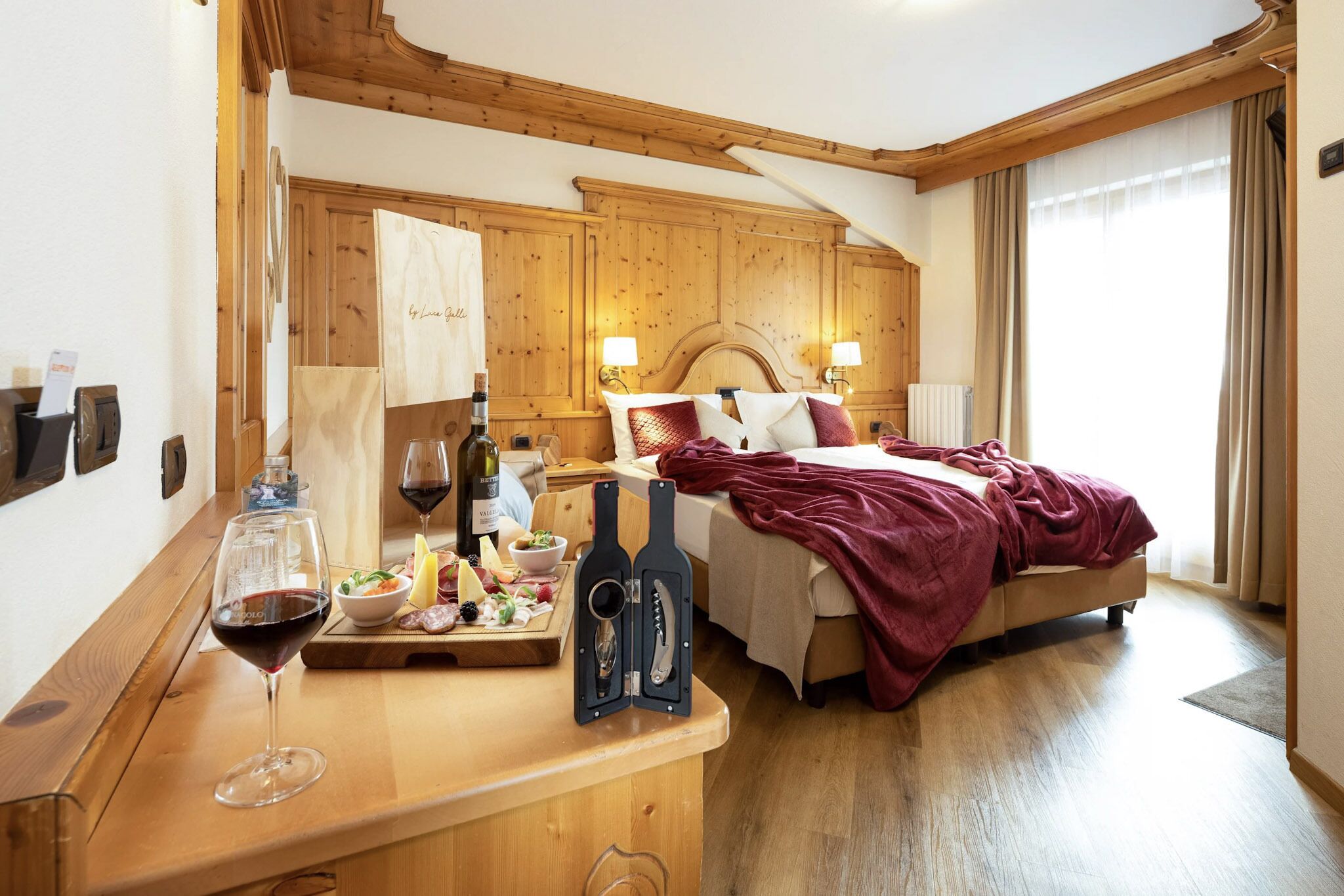 A hotel room with a wooden headboard, a bed with a red blanket, a wooden desk with a wine glass, a bottle of wine, and a bottle opener.