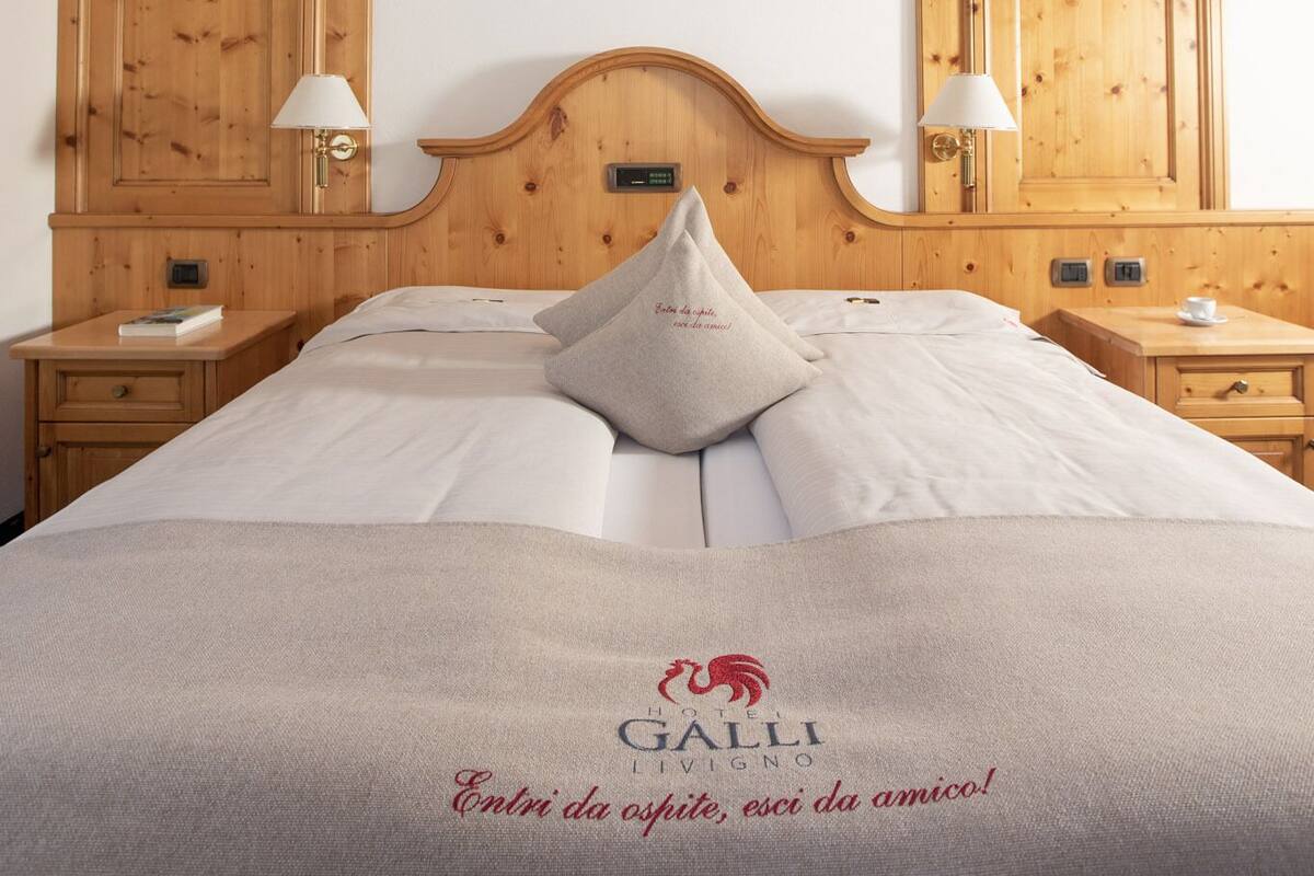 A hotel room with a wooden headboard, two bedside tables with lamps, a bed with a beige blanket, and a heart-shaped decoration on the wall.