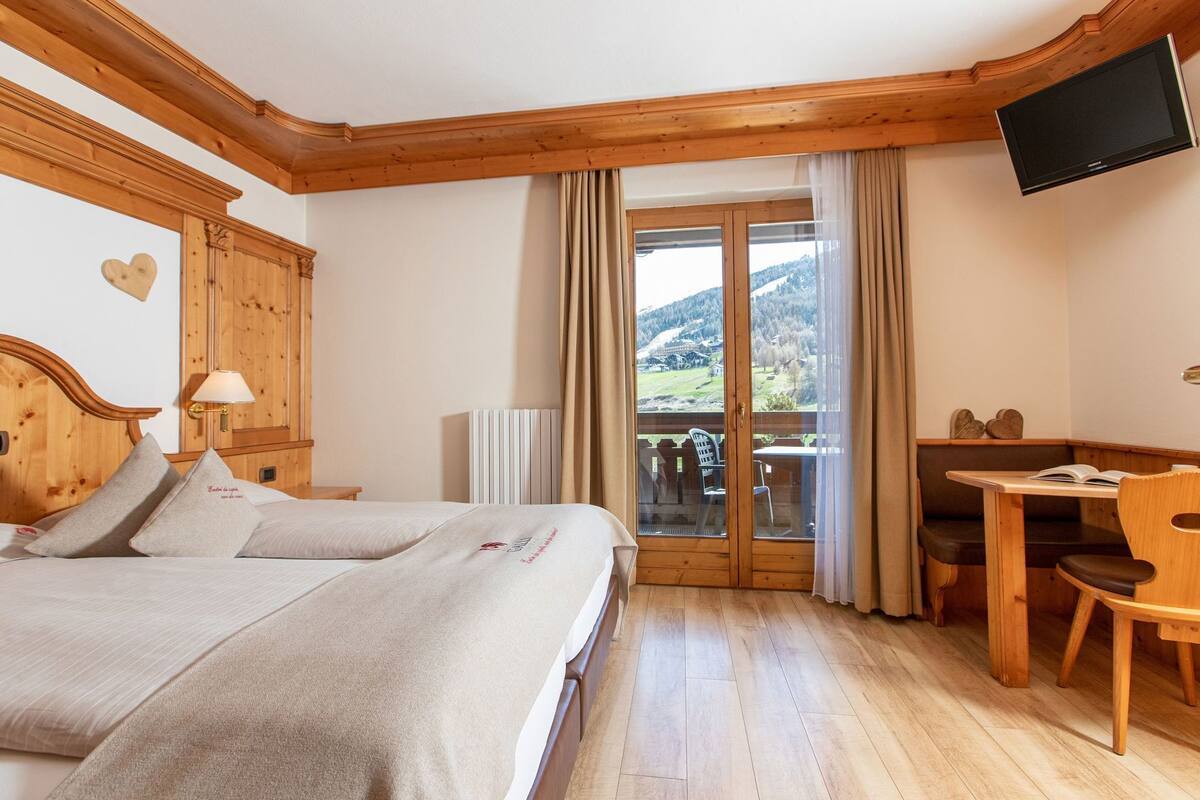 A hotel room with a bed, a desk, a chair, a TV, and a balcony with a view of the countryside.