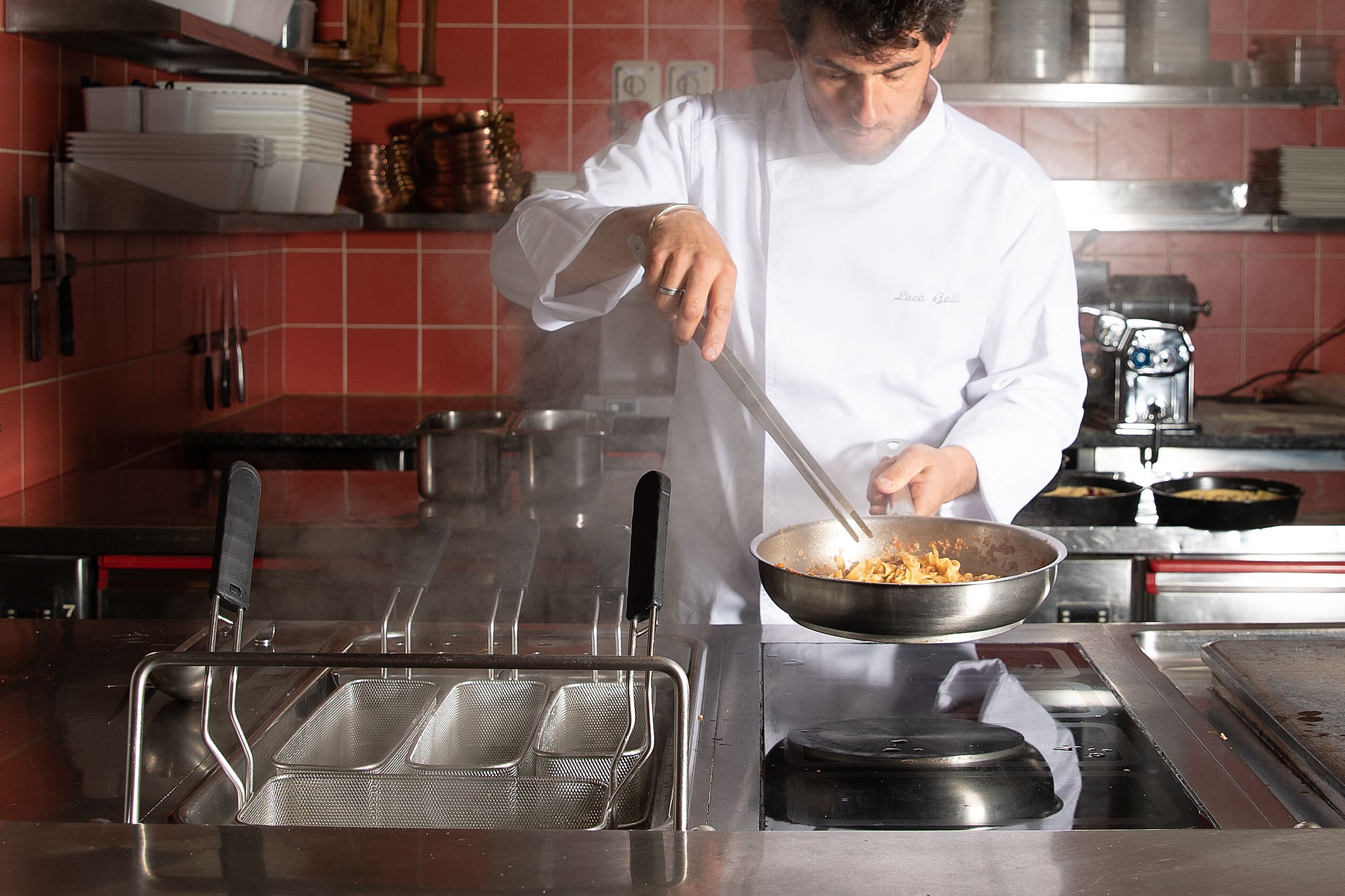 Un chef cuisinant dans une cuisine professionnelle équipée d’appareils en acier inoxydable et dont les murs sont recouverts de carreaux rouges.