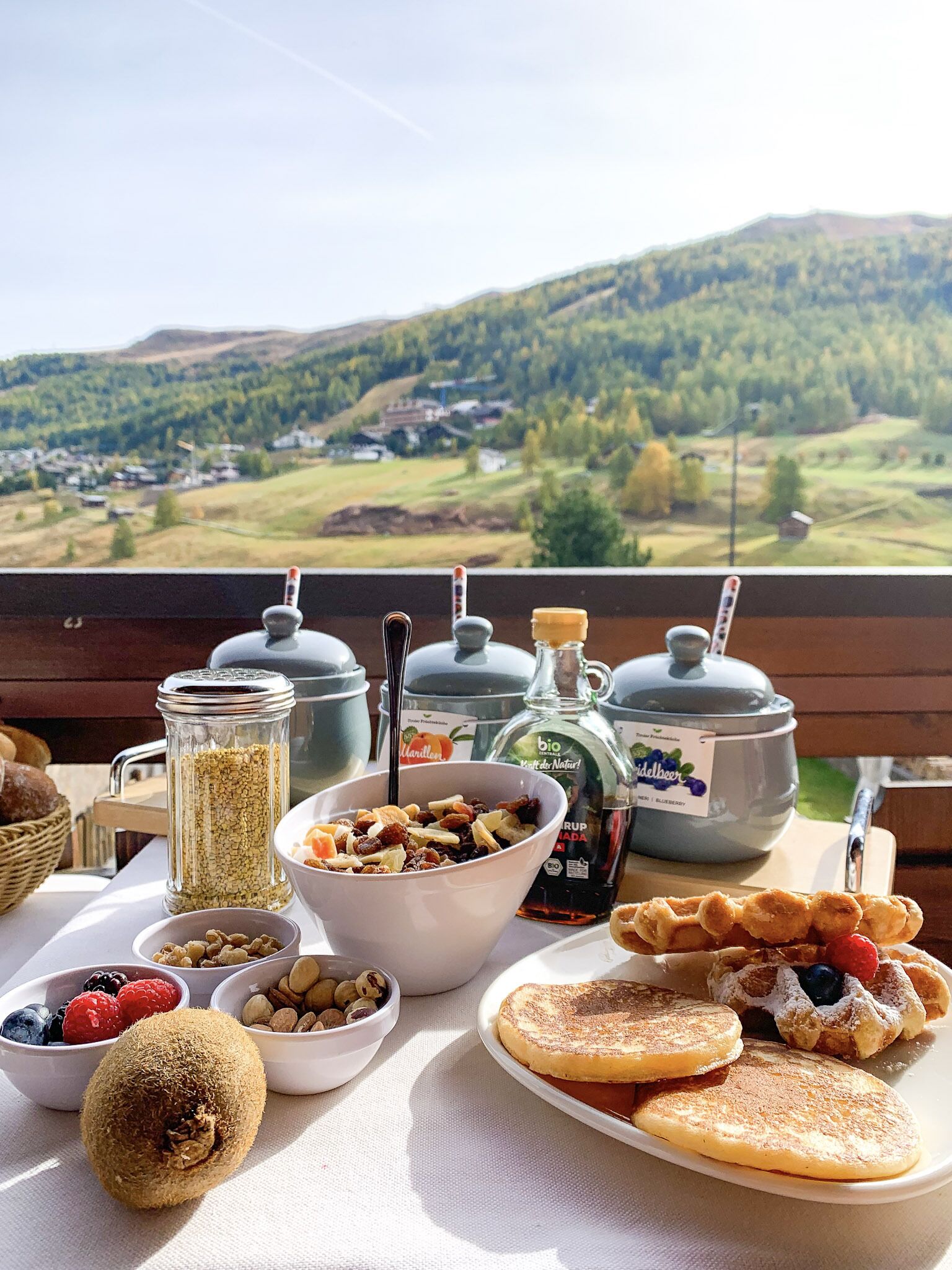 Une table de petit-déjeuner dressée avec divers aliments et condiments, le tout mis en valeur par une vue panoramique sur les montagnes.