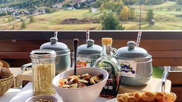 Une table de petit-déjeuner dressée avec divers aliments et condiments, le tout mis en valeur par une vue panoramique sur les montagnes.