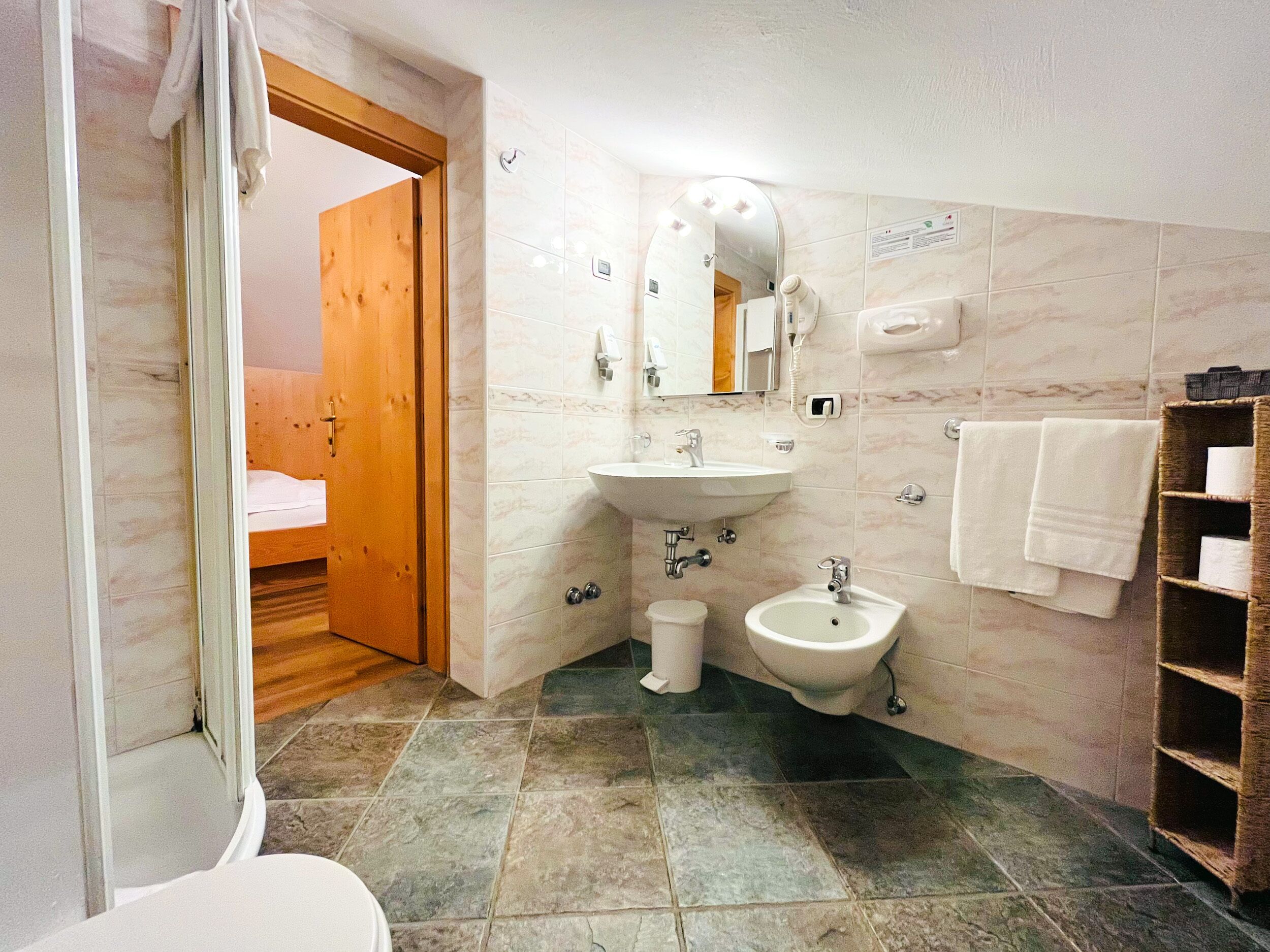 Een badkamer met een wastafel, bidet en een spiegel met verlichting. Er is een houten deur naar een slaapkamer.