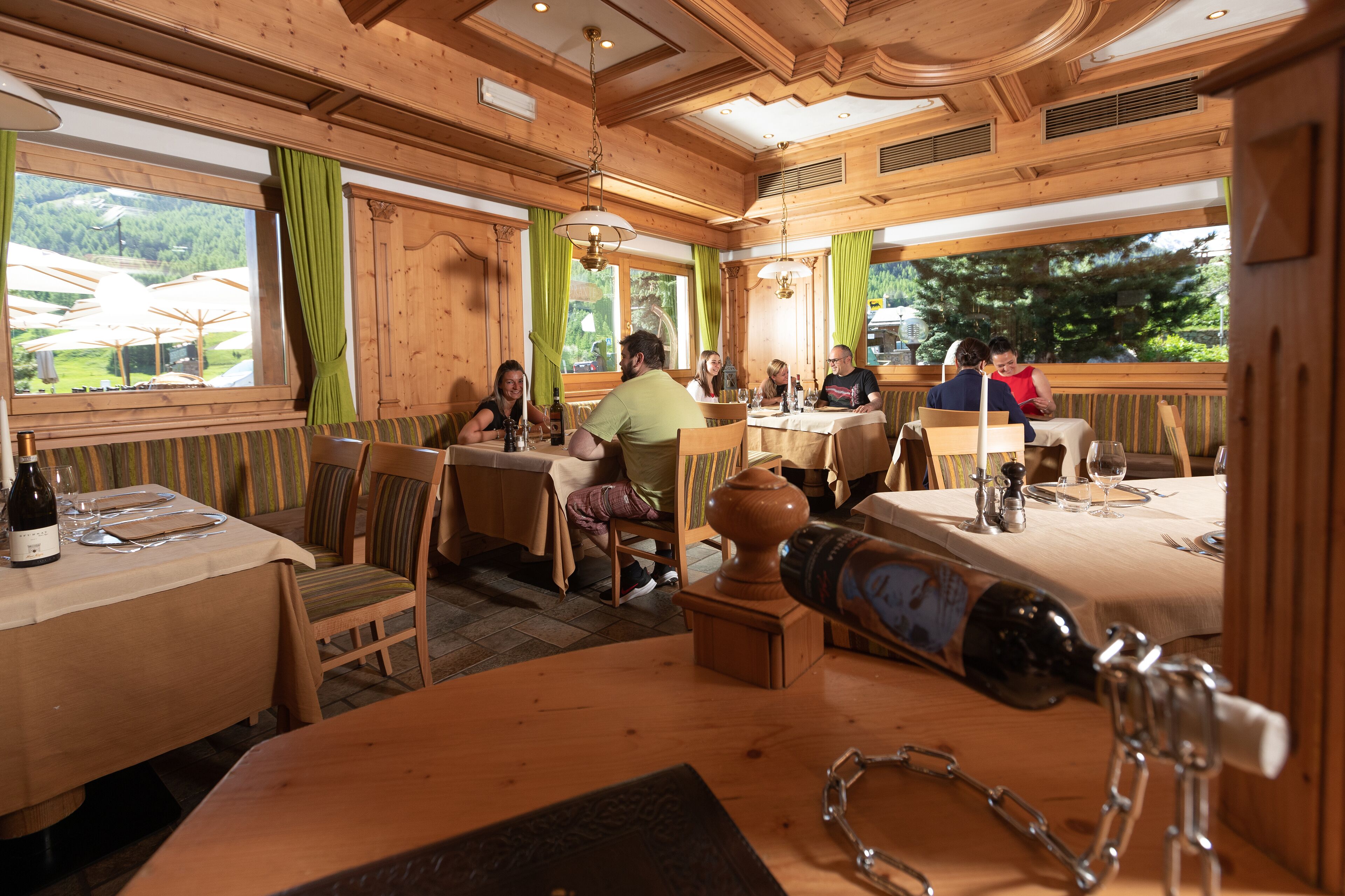 A wooden interior with tables and chairs, a bottle of wine, and a view of greenery outside.