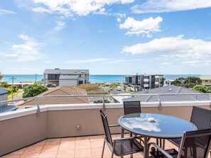 A balcony with a glass table and chairs, overlooking a coastal area with modern buildings and a clear sky. - Boatshed Motel Apartments (Tauranga)