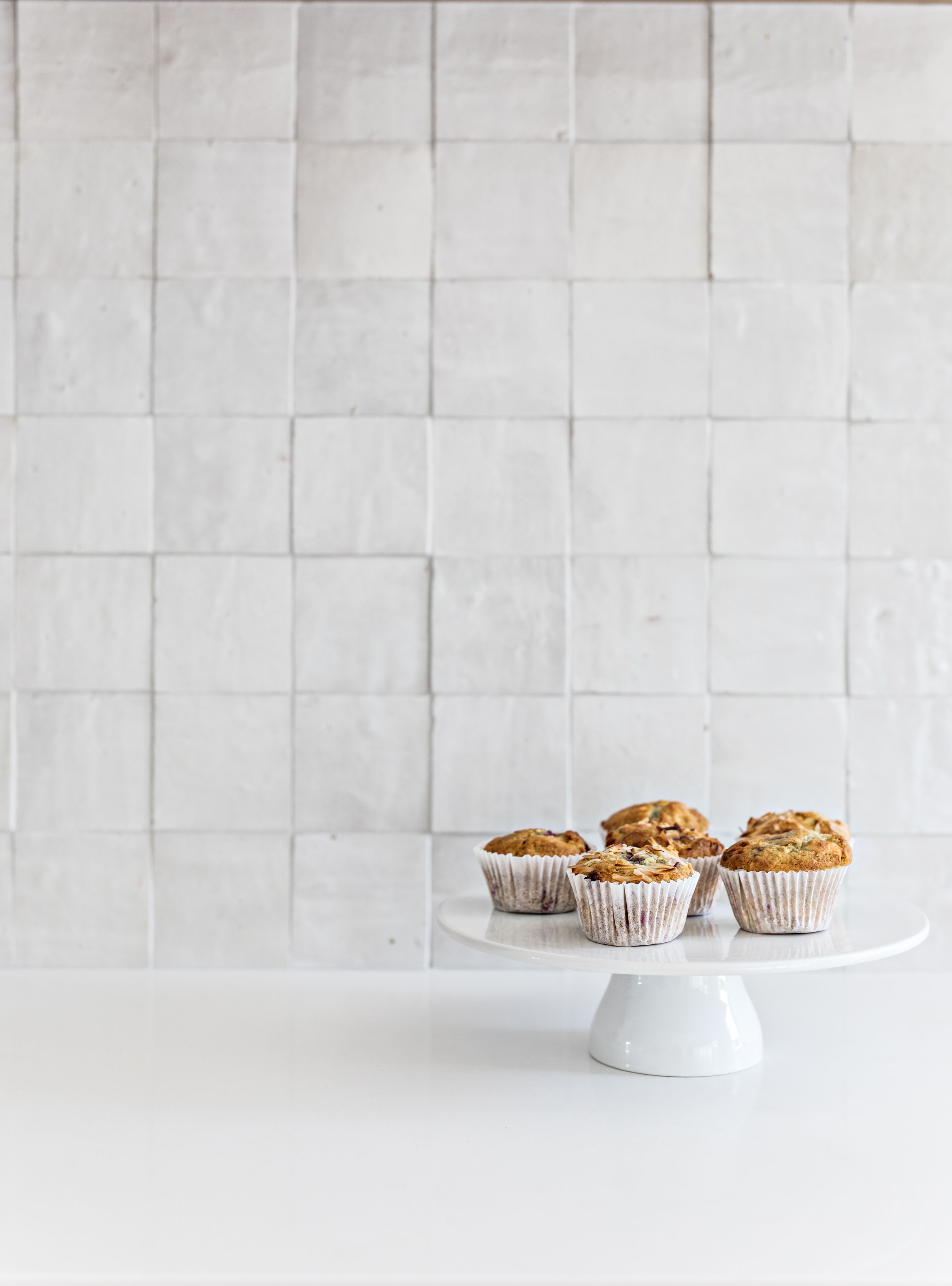 Un mur recouvert de carreaux blancs, sur lequel sont posés trois muffins sur un présentoir à gâteaux blanc.