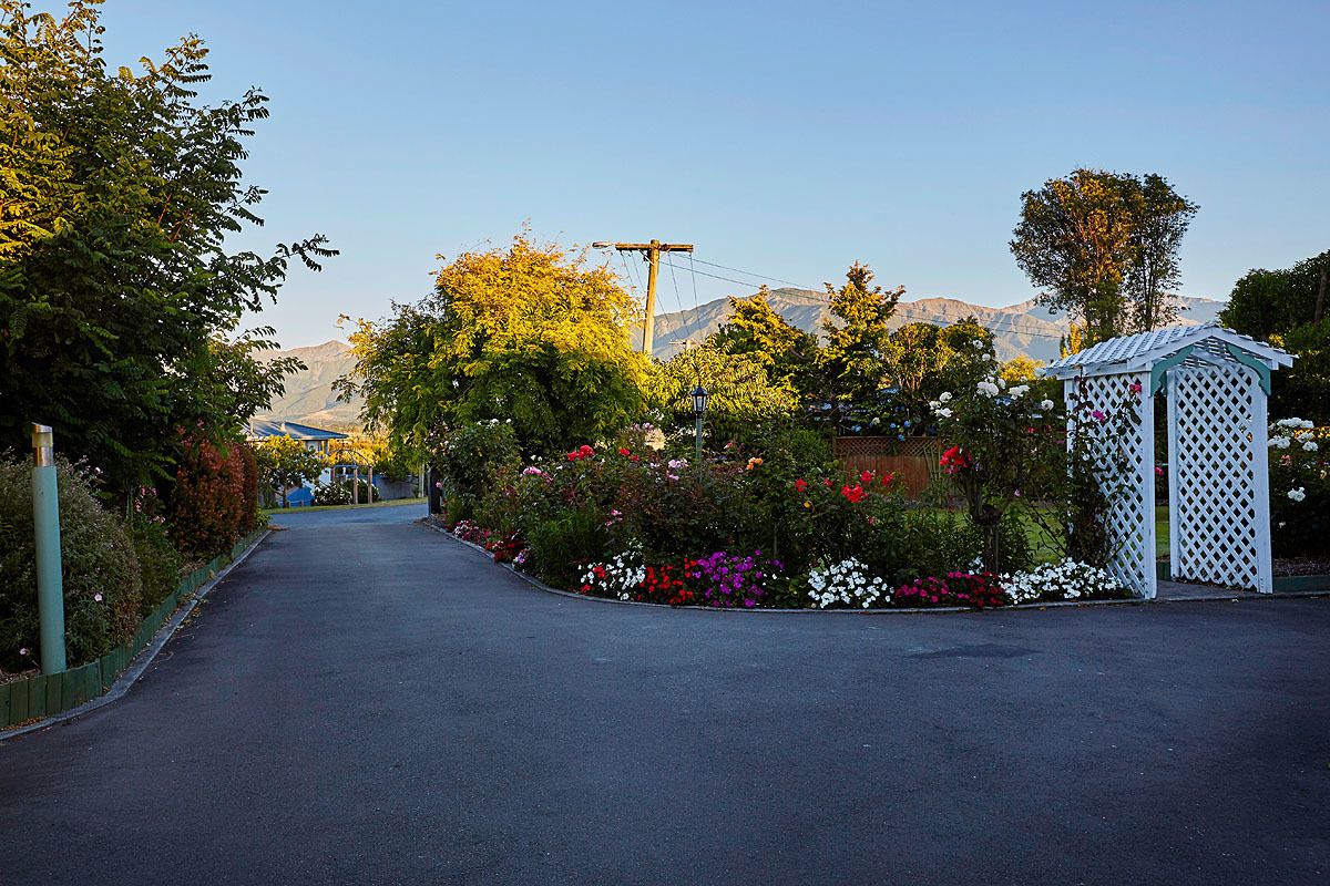 Un camino pavimentado conduce a una zona vallada con una estructura de celosía blanca, rodeada de plantas y árboles florecientes de colores.