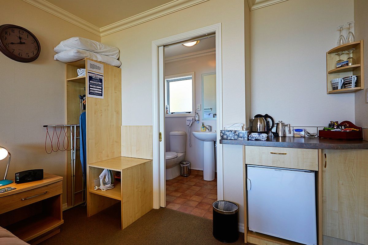 Une chambre d’hôtel compacte comprenant un coin cuisine, une petite salle de bain, une horloge murale et une table de chevet avec une lampe.