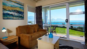 Une pièce avec un canapé marron, une petite table sur laquelle se trouve un vase, une lampe et un tableau accroché au mur. Une grande fenêtre offre une vue sur la mer et les montagnes.