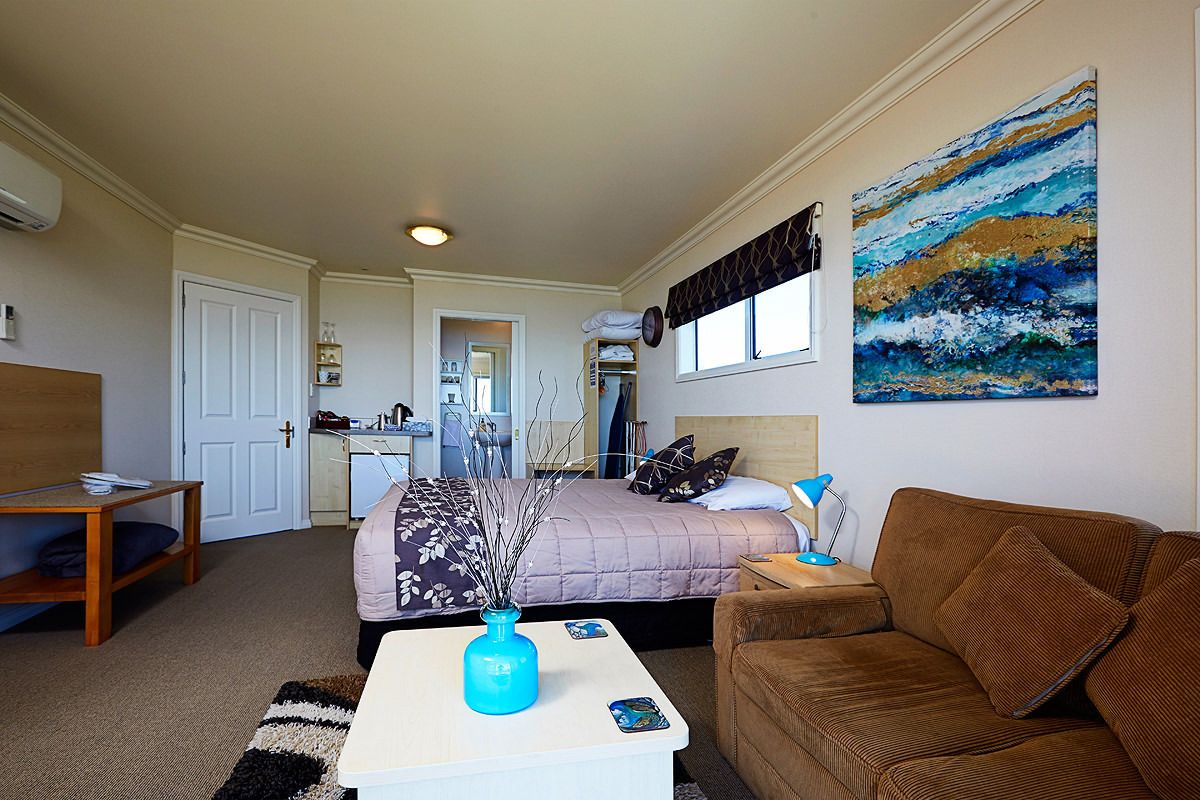 A compact bedroom with a bed, a small table with a vase, a brown sofa, and a painting on the wall.
