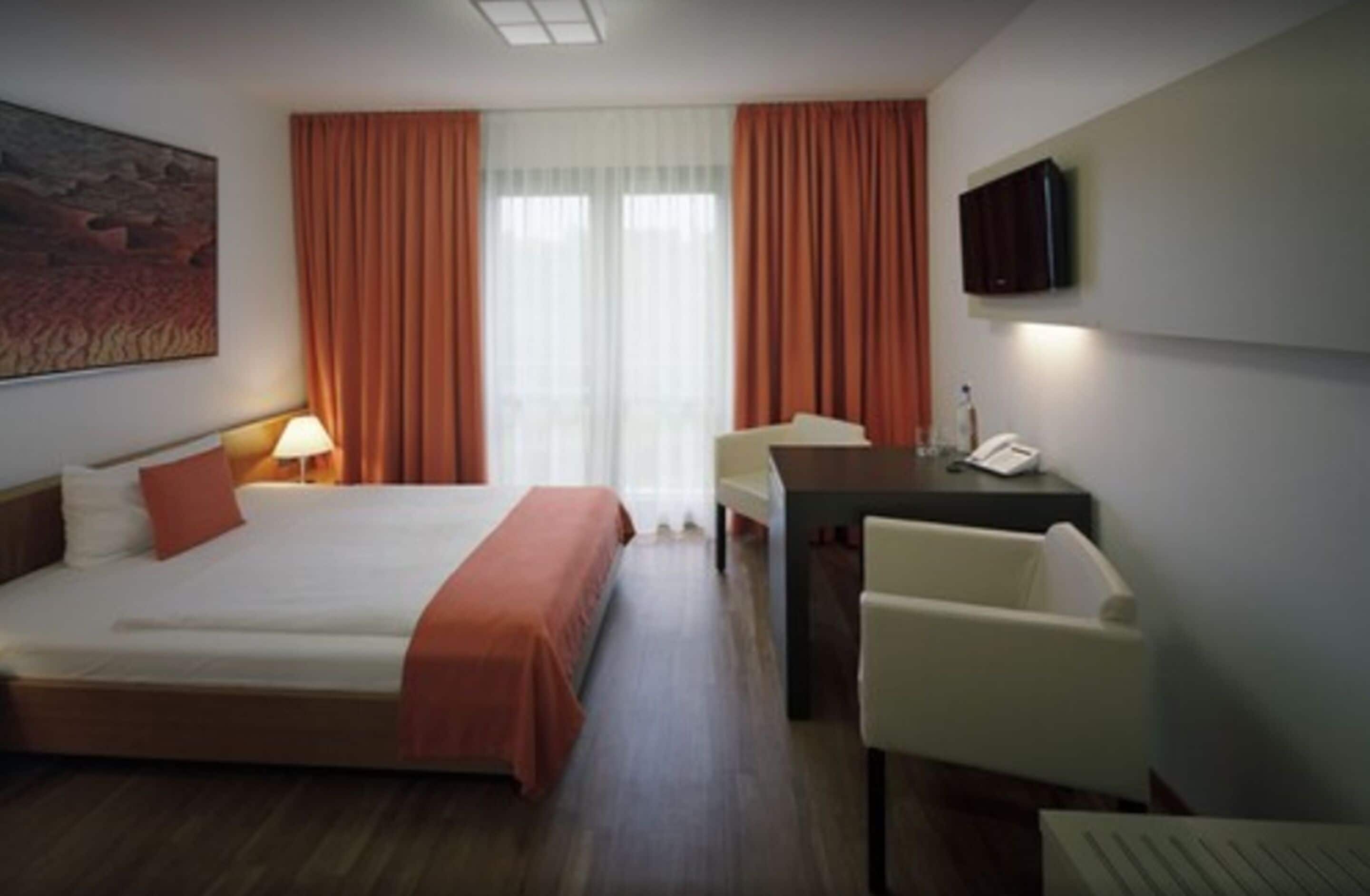 A hotel room with a bed, a desk, a chair, a TV, and a painting on the wall.