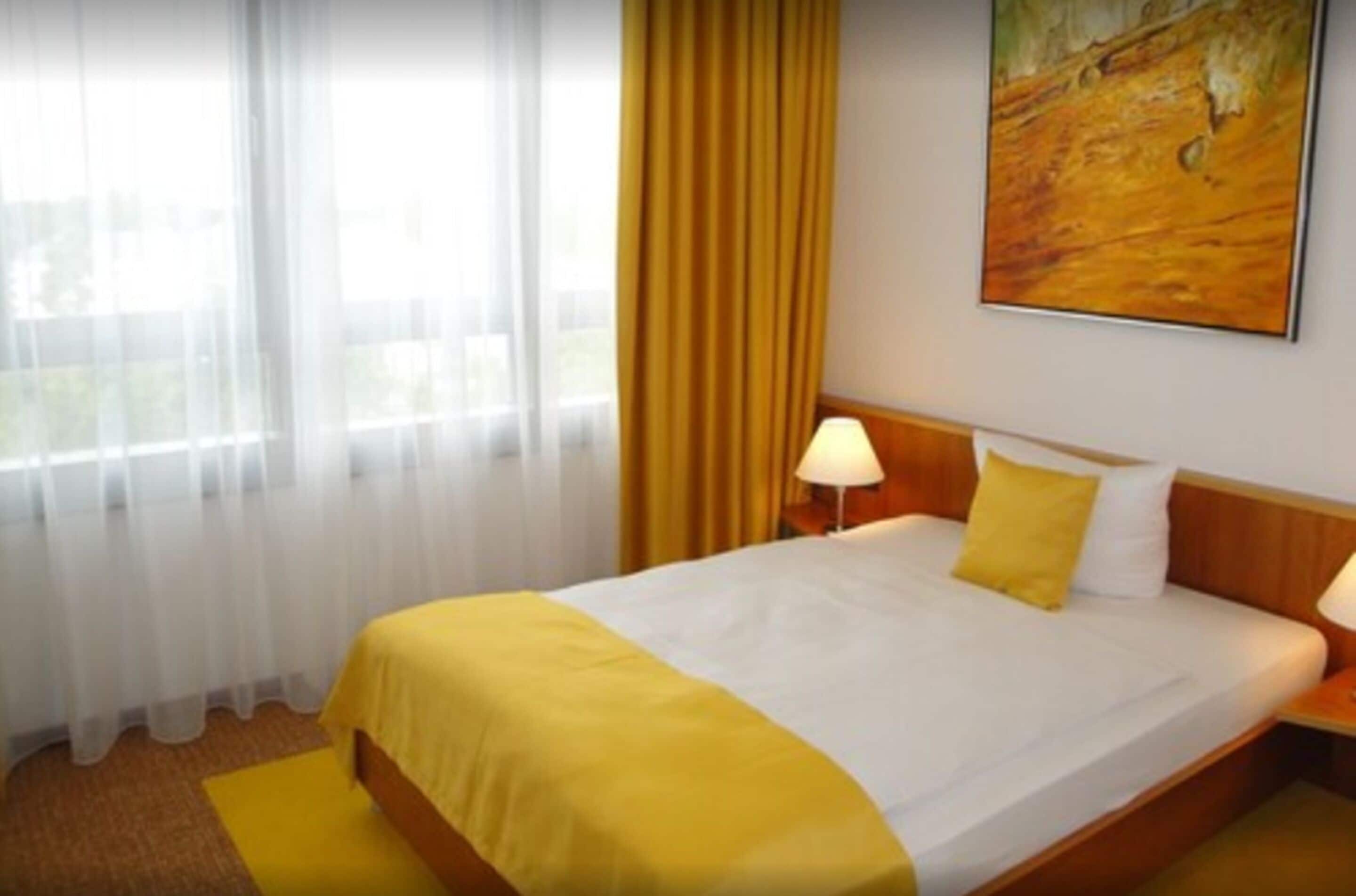 Una camera d'albergo con un letto, un comodino, una lampada, un quadro appeso al muro e una finestra con tende.