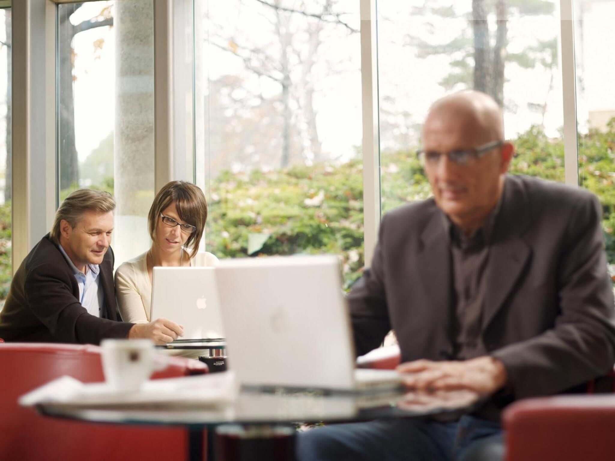 Three people seated at a table with laptops, in a room with large windows and red chairs.