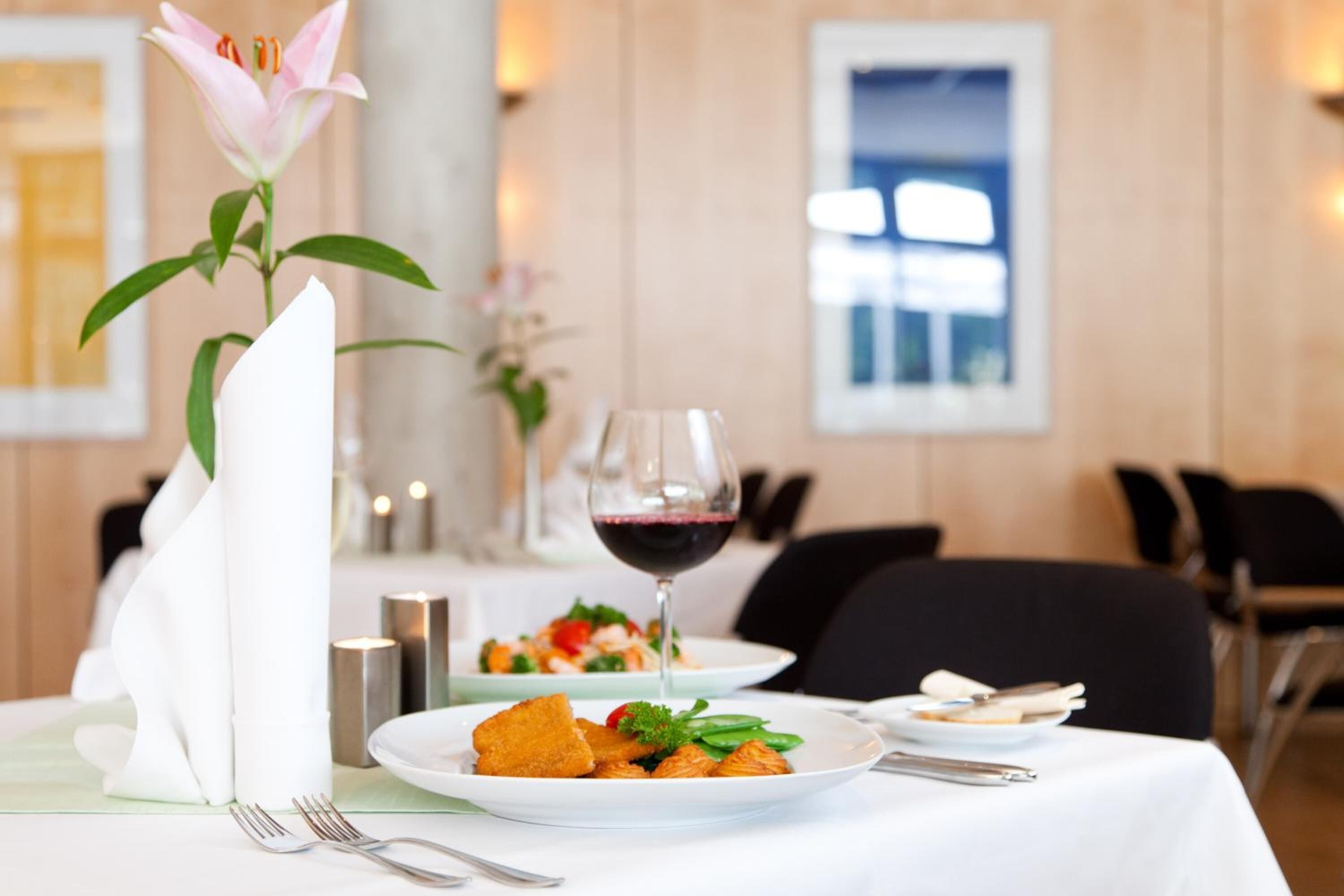 Una mesa de comedor dispuesta con platos de comida, una copa de vino y un arreglo floral.