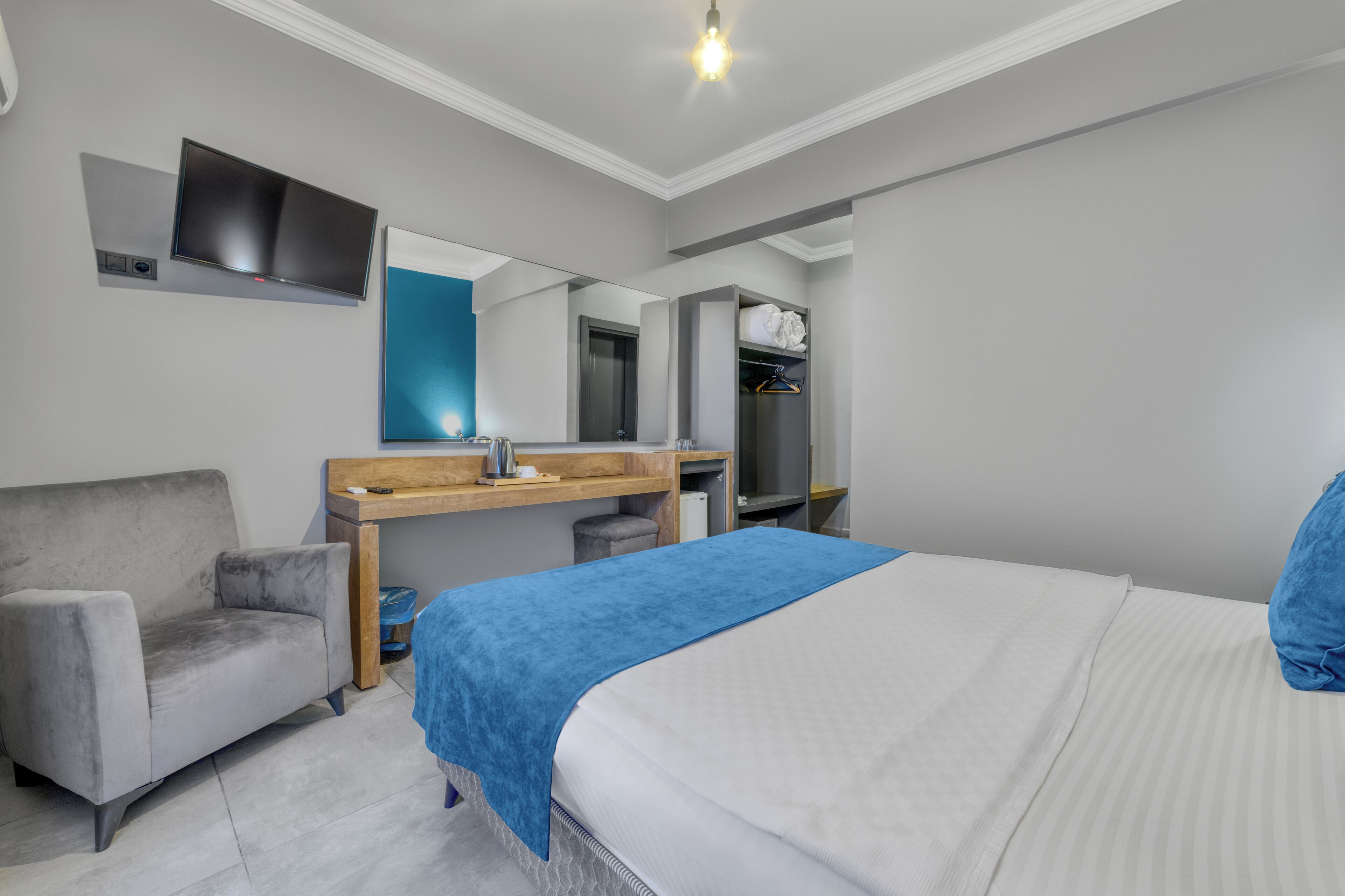 A hotel room with a bed, a gray armchair, a wooden desk with a mirror, and a wardrobe.