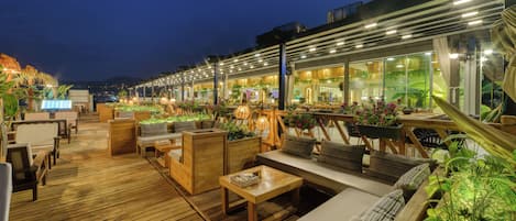 Terraza de madera con mobiliario exterior, incluyendo sofás y mesas, y una zona de restaurante con ventanas amplias y plantas de interior.