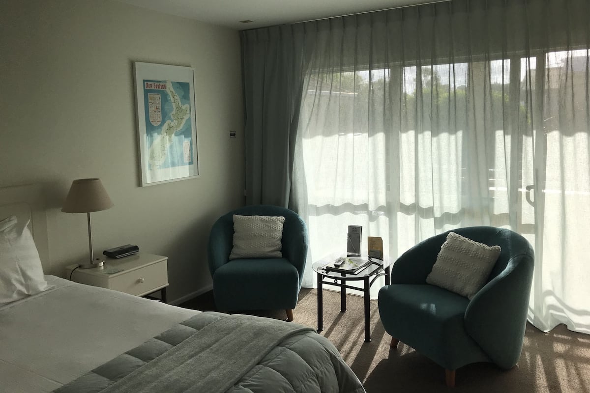 A hotel room with a bed, two armchairs, a nightstand, a lamp, a map on the wall, and a window with sheer curtains.