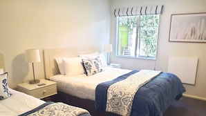 A hotel room with two beds, a window with a view, a bedside table with a lamp, and a wall-mounted artwork. - Tuscana Motor Lodge (Christchurch)
