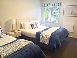 A hotel room with two beds, a window with a view, a bedside table with a lamp, and a wall-mounted artwork. - Tuscana Motor Lodge (Christchurch)