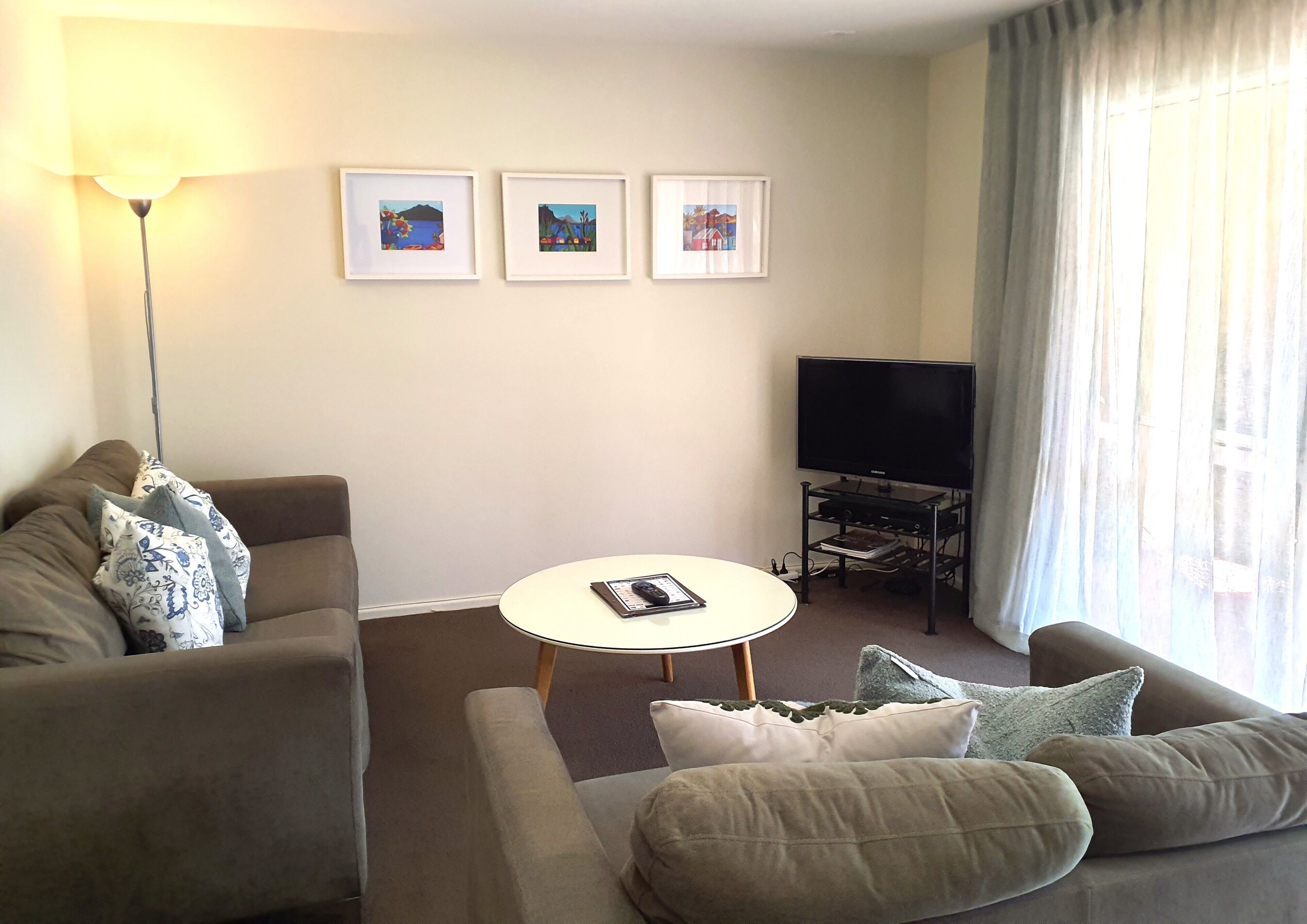 Uma sala de estar com um sofá, uma mesinha redonda, uma TV em um rack e três quadros na parede.