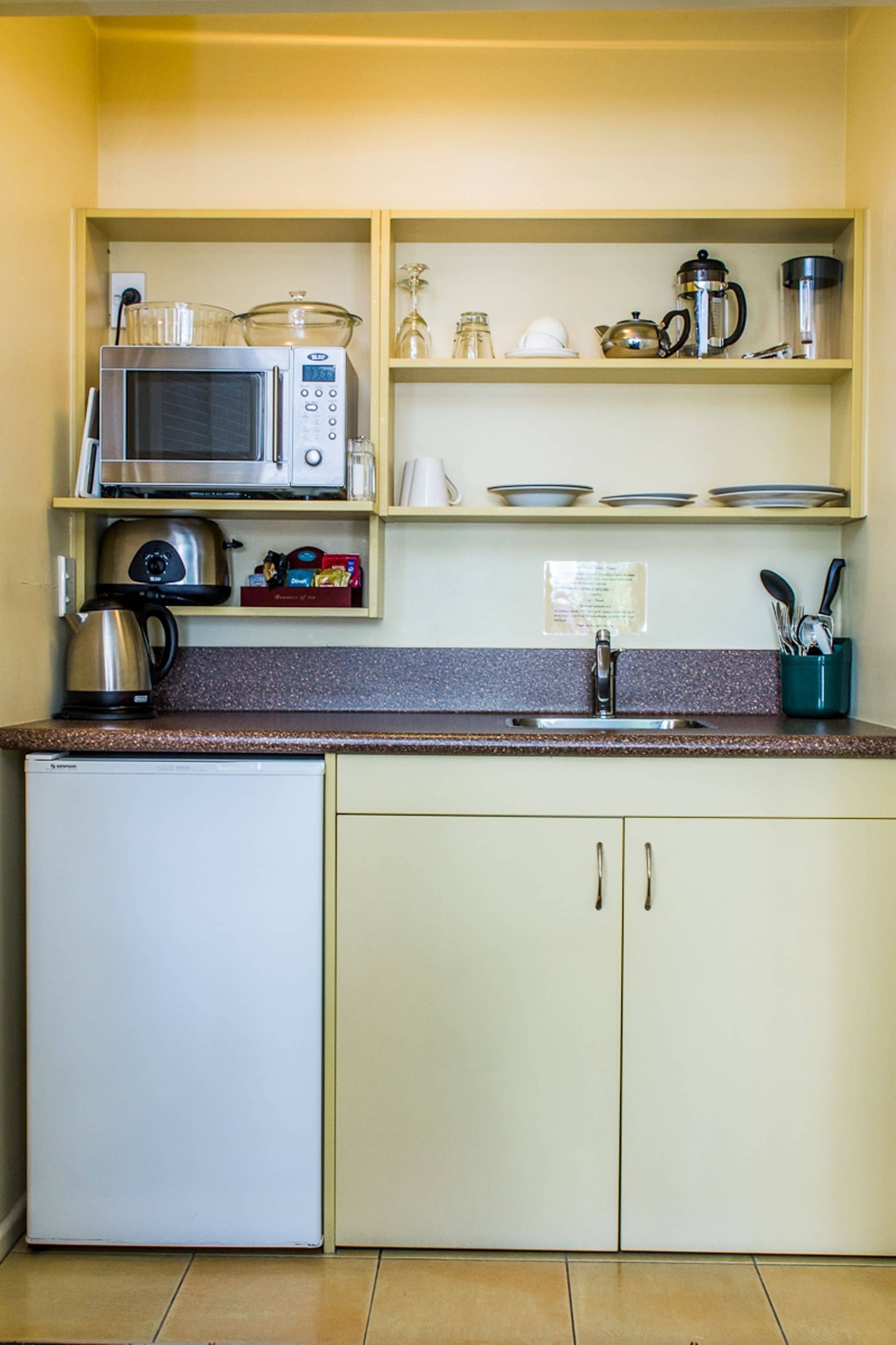 A compact kitchen with a microwave, kettle, and coffee maker on the counter, and a refrigerator below.