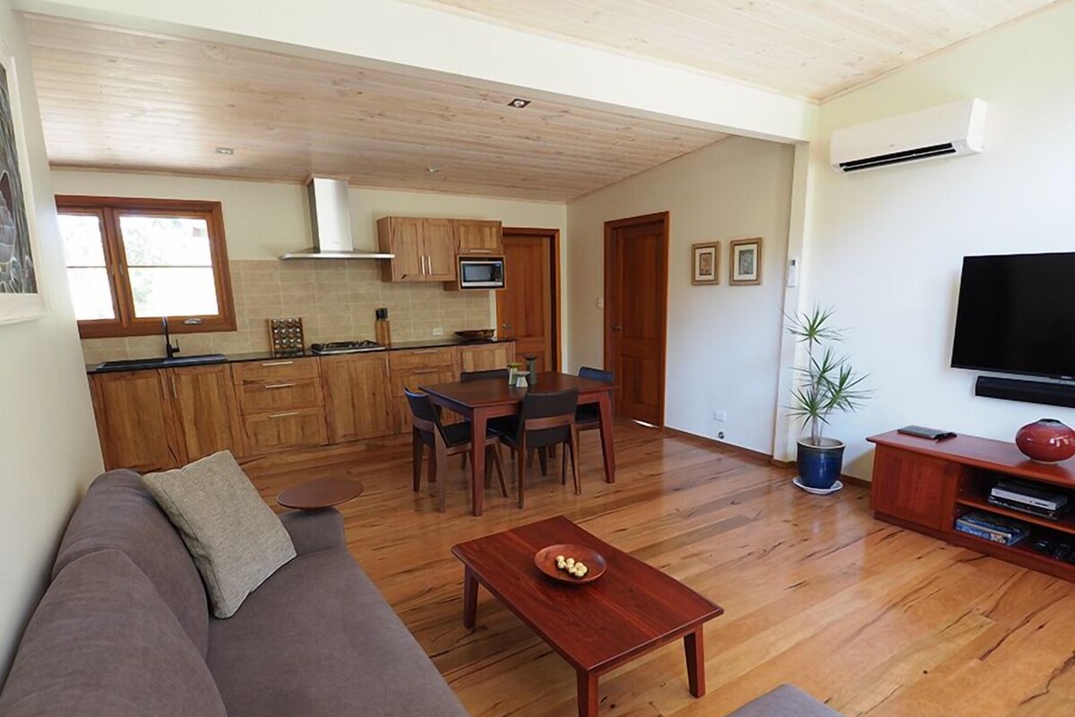 Ein modernes Wohnzimmer mit Holzboden, einem braunen Sofa, einem Esstisch mit Stühlen, einem Fernseher und einer Küchenzeile mit Holzschränken und einer Mikrowelle.