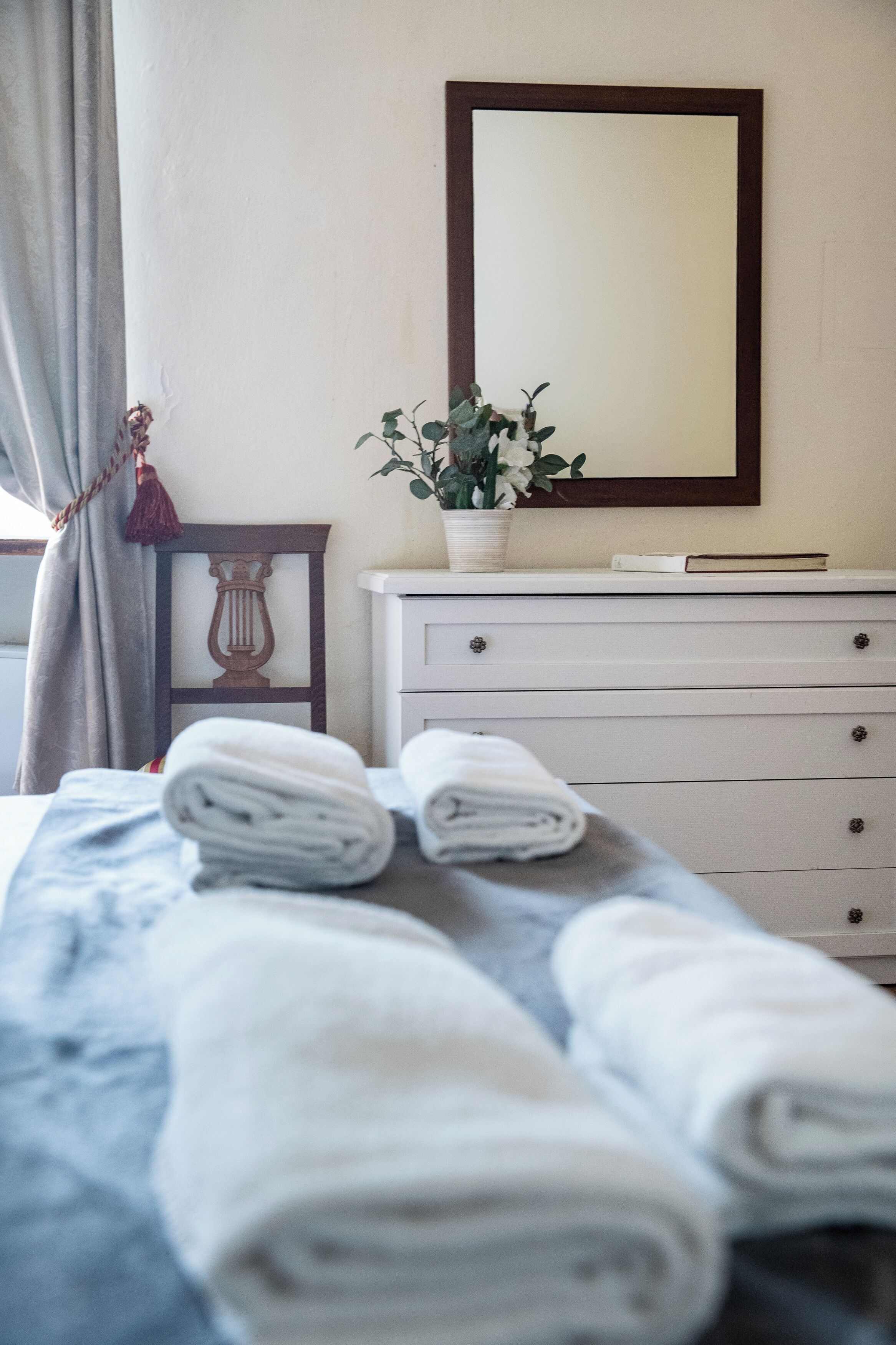 A neatly made bed with rolled towels, a bedside table with a book and a potted plant, and a mirror on the wall.