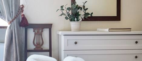 A neatly made bed with rolled towels, a bedside table with a book and a potted plant, and a mirror on the wall.
